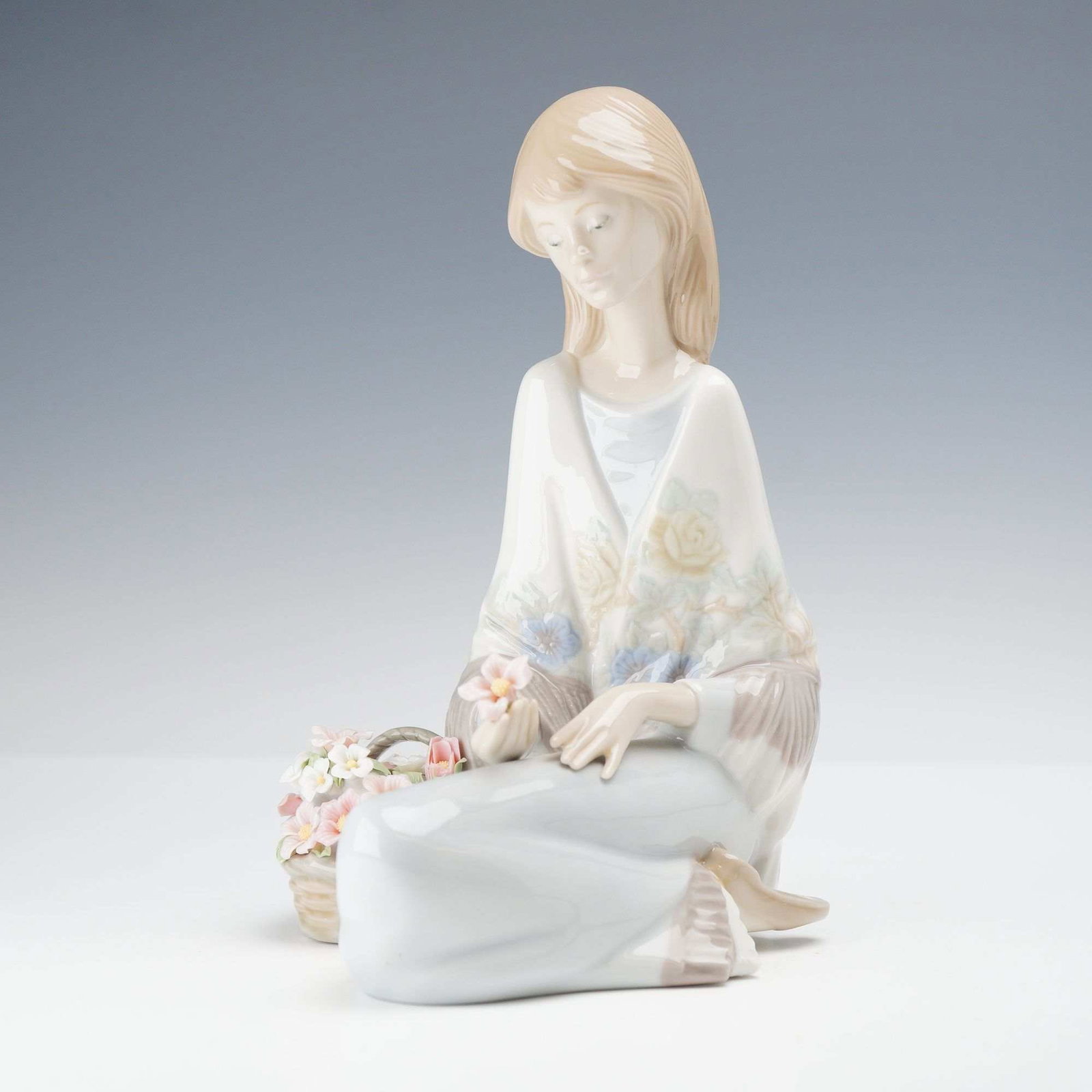 Lladro Porcelain Figurine, Flower Song 01007607: The piece depicts a young woman kneeling in a contemplative pose, dressed in a pale wrap decorated with soft blue and yellow floral motifs. She holds a single delicate flower in her hand, while a wove