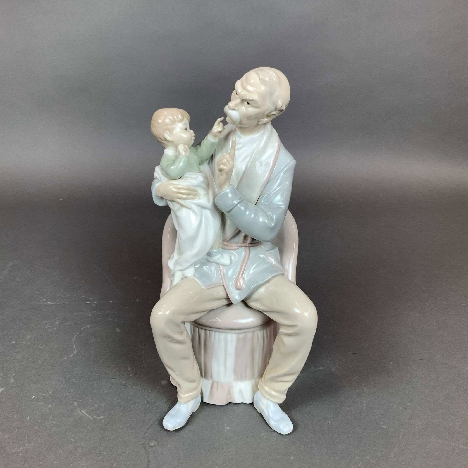 Lladro Porcelain Figurine, The Grandfather 01004654: A Lladro glazed porcelain figurine titled The Grandfather, model number 01004654. The composition depicts a grandfather seated on a draped stool wearing a pale blue robe over tan trousers and white sl