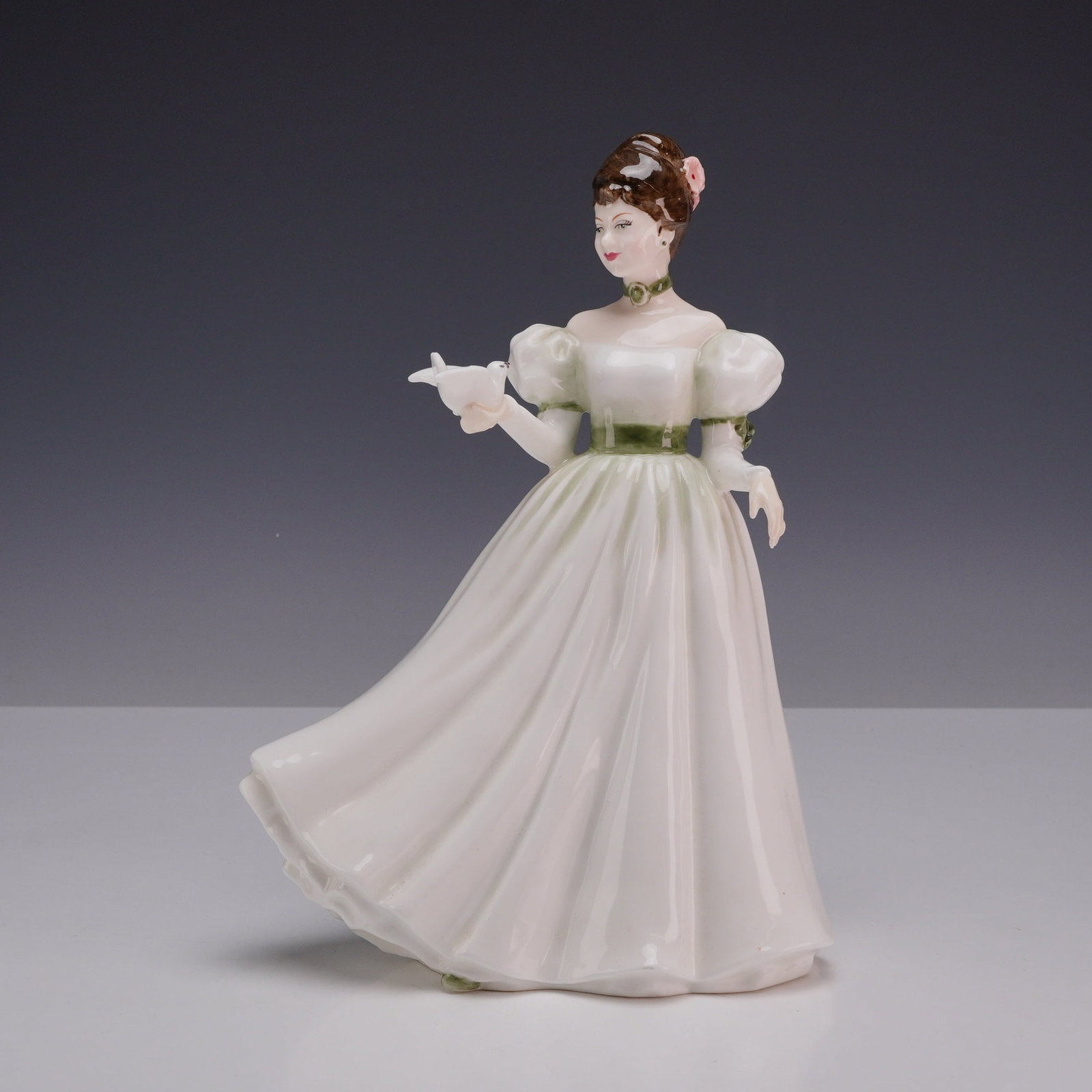 Royal Doulton Bone China Figurine, Brianna HN4126: Depicts a young woman in an off-white or cream-colored gown with puffed sleeves and green accents at the neckline, waist, and sleeves. She is posed holding a small white bird in her right hand, and he