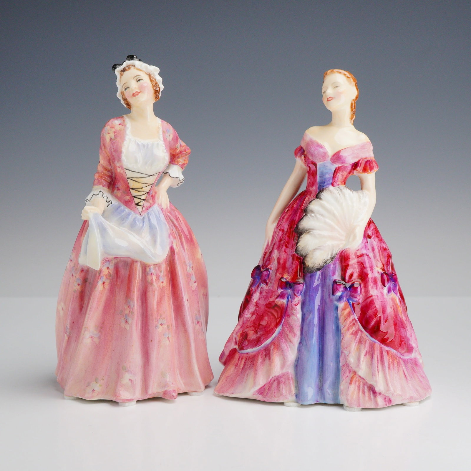 2pc Royal Doulton Bone China Figurines Mary Jane HN 1990 and Elfreda HN 2078: A group of hand-painted glazed bone china figurines produced by Royal Doulton in England. The first figurine, Mary Jane (model HN 1990), features an 18th-century style pink floral gown with a white ap