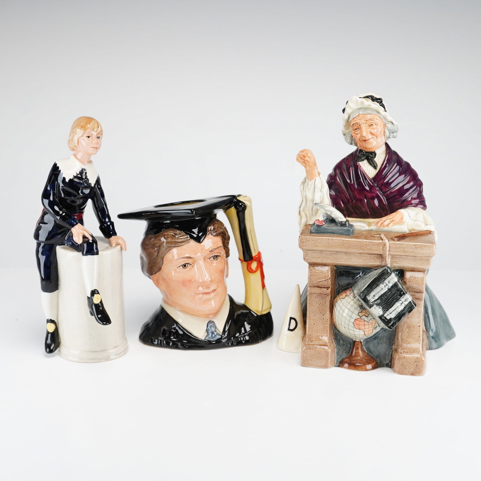 3pc Royal Doulton Figurines and Jug, Schoolmarm, Little Lord Fauntleroy, and The Graduate: Three glazed porcelain pieces by Royal Doulton, England, including two figurines and one character jug. The collection includes The Schoolmarm (HN 2223), depicting an elderly woman seated at a desk wi