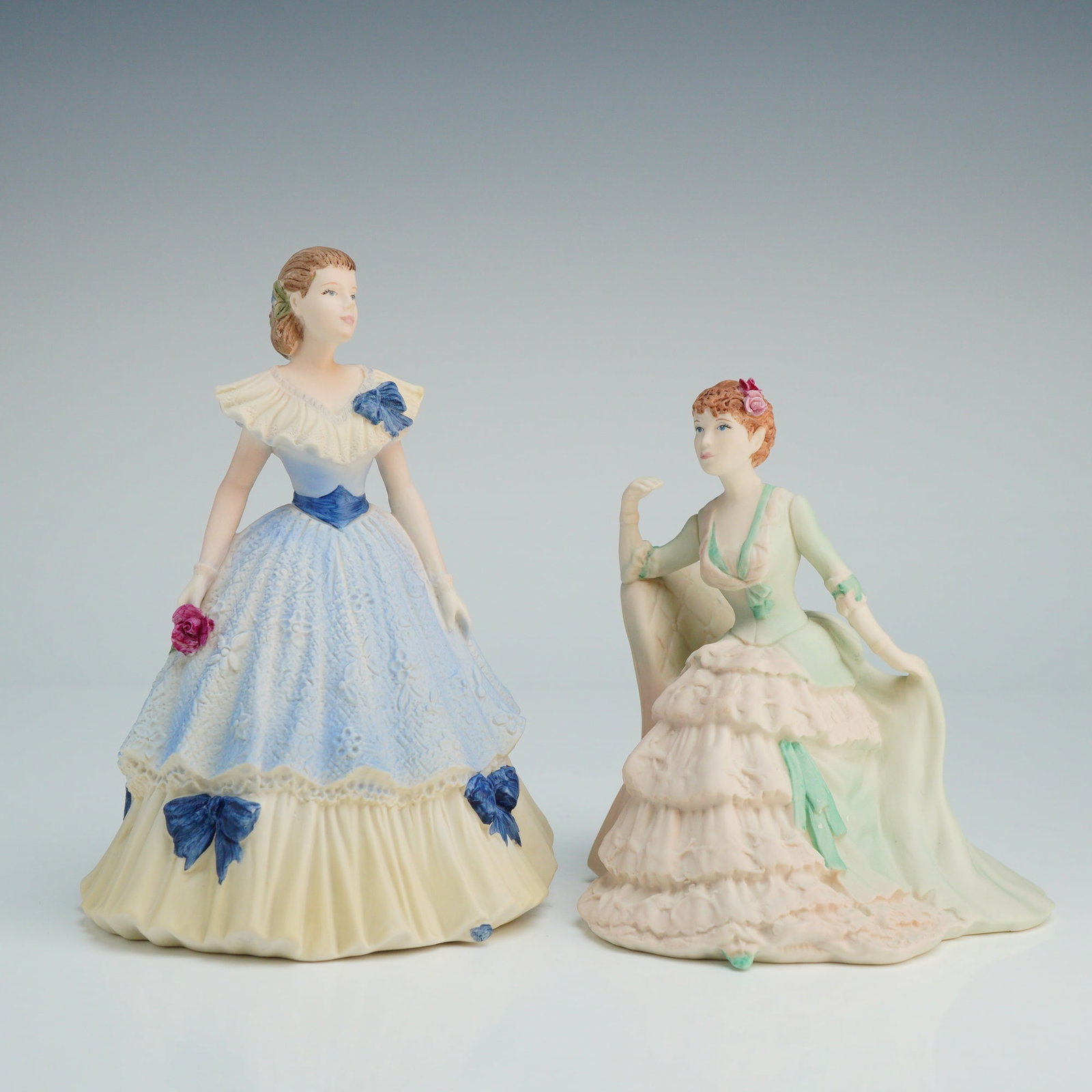2pc Coalport Porcelain Beau Monde Figurines, Clara & Lynne: Both items are hand-decorated with a matte finish. Lynne, is a standing figure wearing a ruffled blue and cream gown with dark blue bows and floral hair details, holding a single pink rose. Clara depi