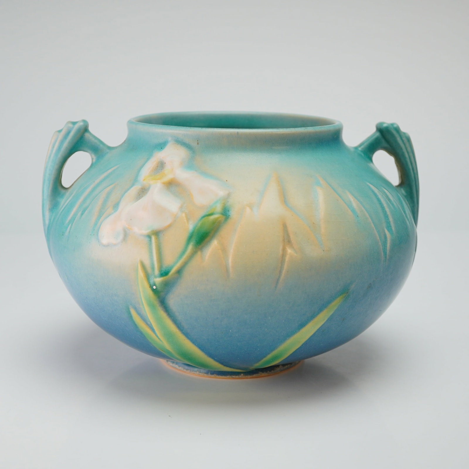 Roseville Pottery Blue Two Handled Jardiniere, Iris: A Roseville Art Pottery Iris pattern two-handled squat vessel, circa 1939. The ceramic vessel features a bulbous form with a matte glaze transitioning from a light teal at the rim to a deep blue base.
