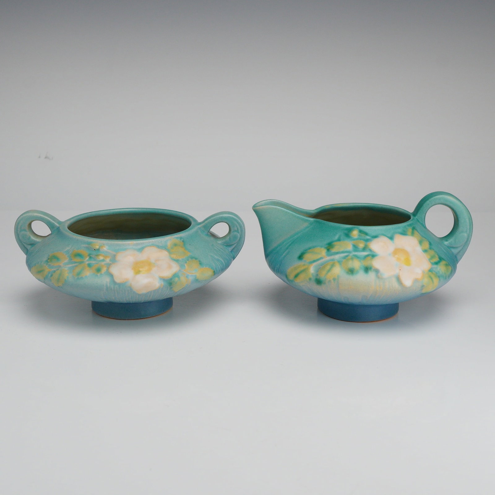 Roseville Pottery Blue Creamer and Sugar Bowl Set, White Rose: A two-piece set of American art pottery from Roseville Pottery of Zanesville, Ohio, in the White Rose pattern, introduced circa 1940. The set includes a creamer and a twin-handled open sugar bowl, bot