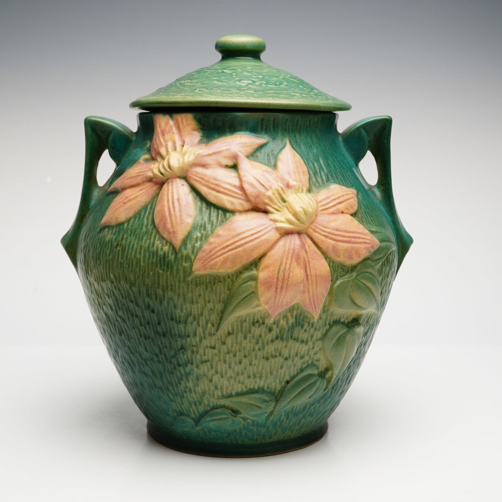 Roseville Pottery Lidded Cookie Jar, Clematis: This mid-century American art pottery piece features a bulbous, tapering body with two small integrated handles and a domed lid with a central knob finial. The decoration is relief-molded with hand-ap