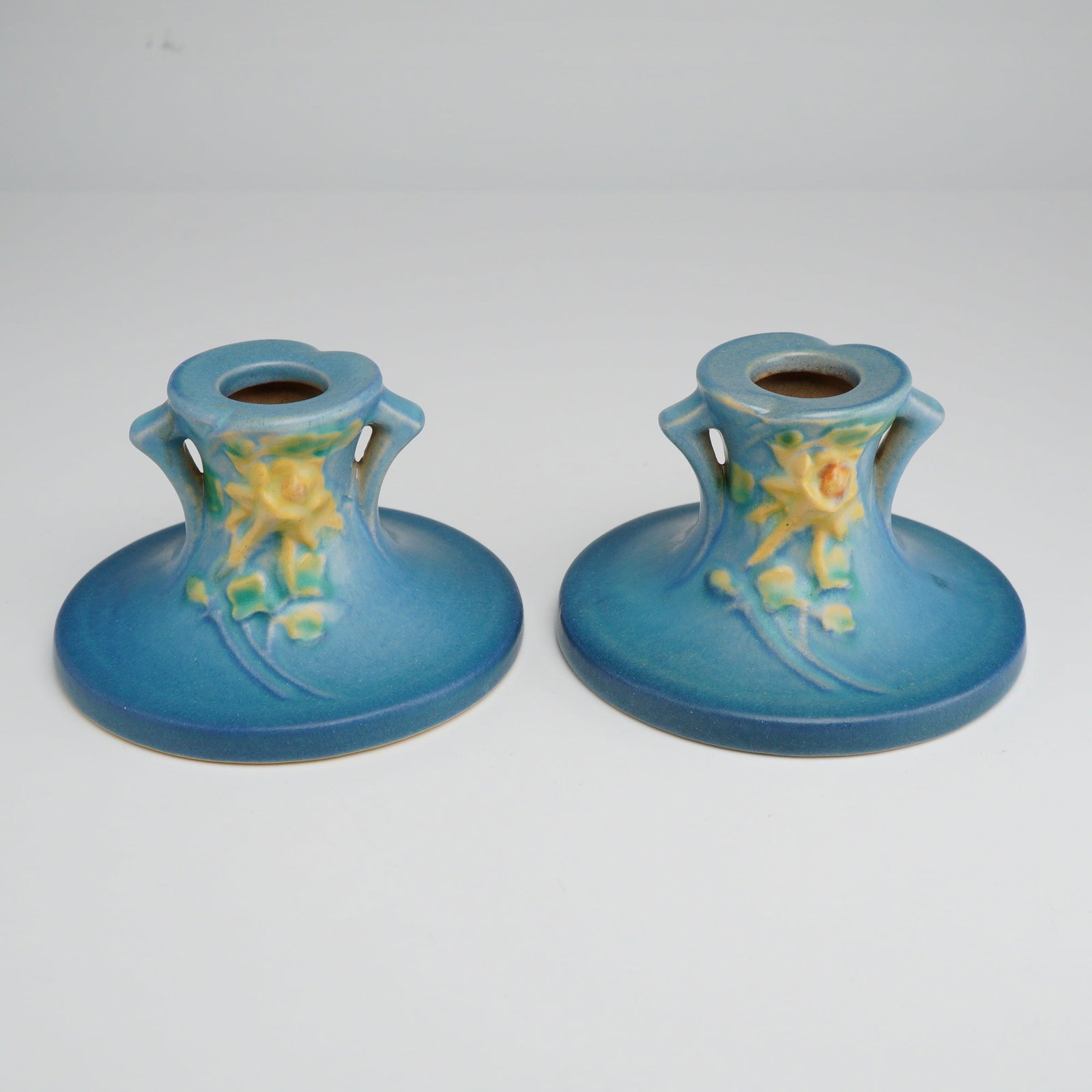 Pair of Roseville Pottery Bermuda Blue Candleholders, Columbine: These pieces feature the Bermuda Blue matte glaze, characterized by a soft teal ground with embossed yellow lily blossoms and green foliage in relief. The form consists of a broad, flared circular bas