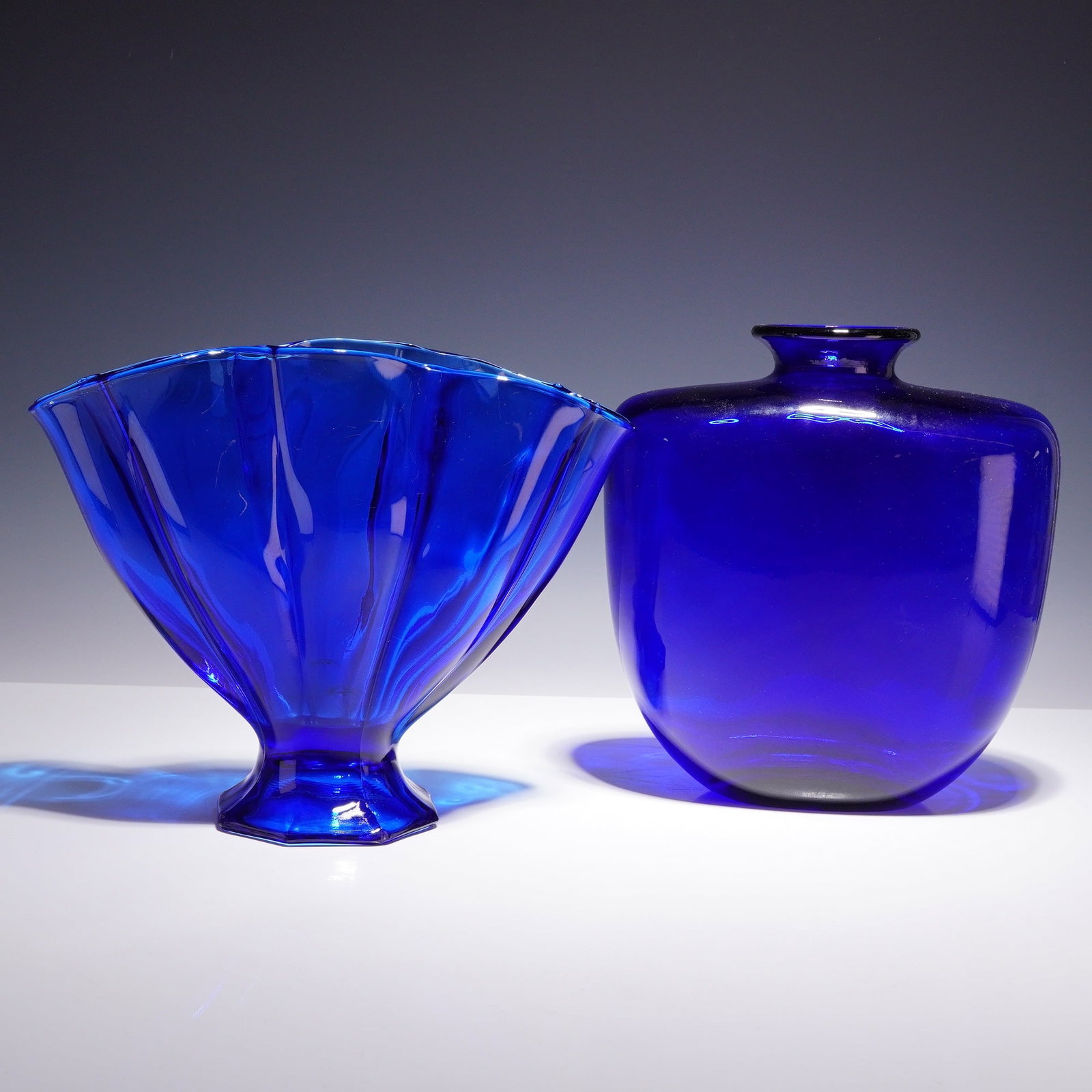 Two Cobalt Blue Glass Vases: This lot consists of two decorative vases in deep cobalt blue glass. The first item is a pressed glass vase featuring a flared, ruffled rim and a vertically ribbed body that tapers down to a short ped