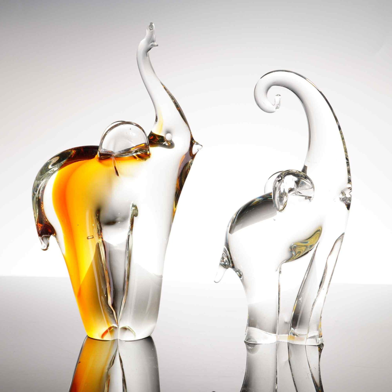 2pc Art Glass Elephant Figurines: These hand-blown elephant figurines feature a stylized, minimalist design characteristic of mid-to-late 20th-century Venetian art glass. The larger figurine is accented with internal swirls of amber a