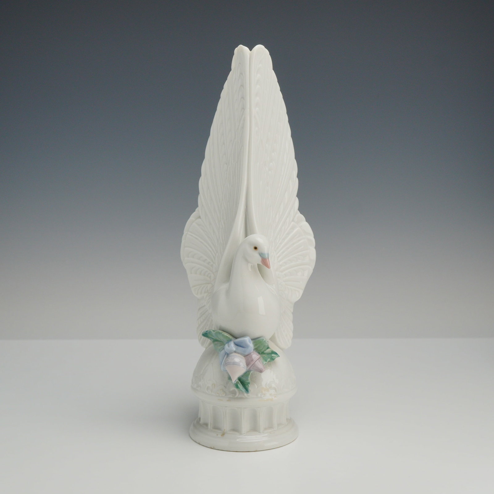 Lladro Porcelain Tree Topper Figurine, Messages of Love 6643: This serene porcelain dove is depicted with expansive wings spread wide and tail feathers elegantly fanned as it perches atop a circular pedestal. Sculptor Francisco Polope designed this graceful figu