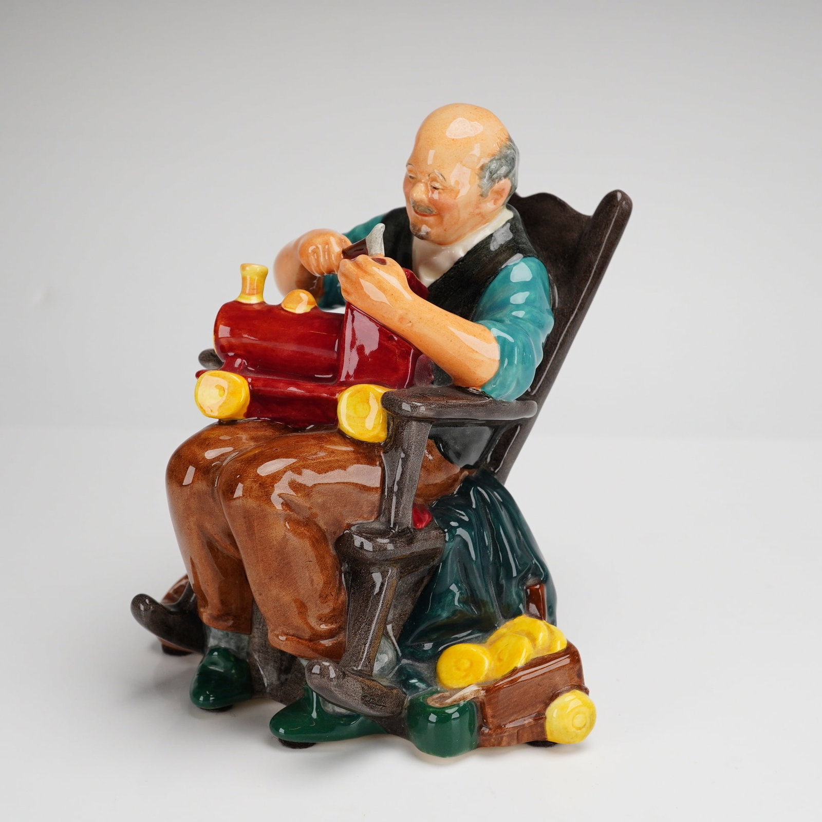 Royal Doulton Figurine The Toymaker HN2250: This Royal Doulton figurine, titled The Toymaker, is a hand-painted porcelain model identified as HN2250. The piece depicts an elderly man in a teal shirt and brown trousers seated in a dark brown roc