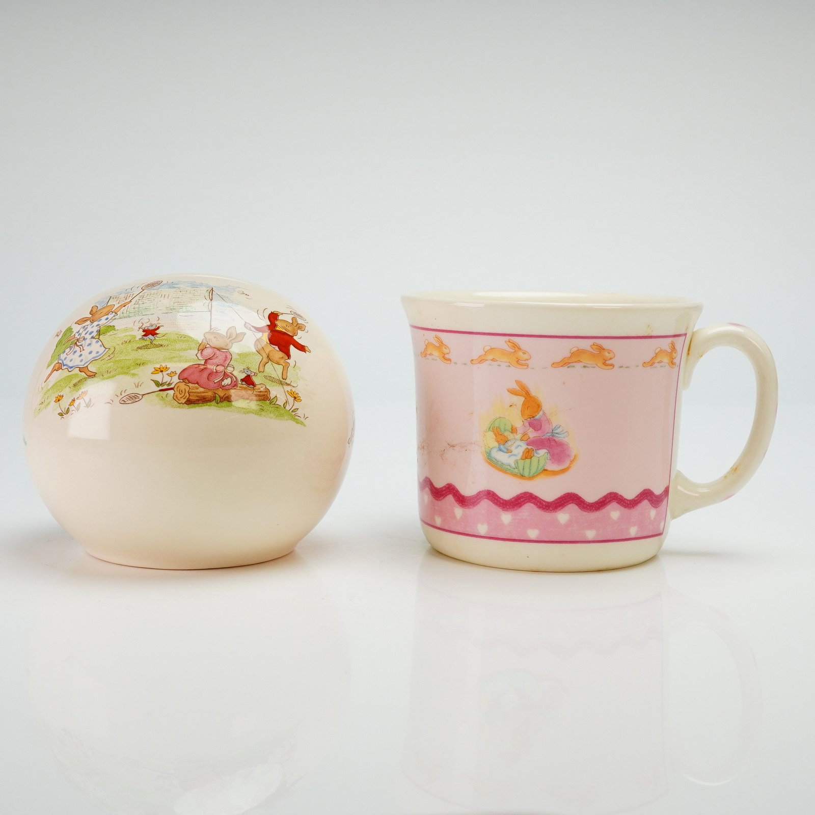 2pc Royal Doulton Bunnykins Bone China Money Ball & Mug: This 2pc Royal Doulton Bunnykins bone china set consists of a spherical money ball and a single-handled mug. The money ball is decorated with a tennis match scene featuring anthropomorphic rabbits and