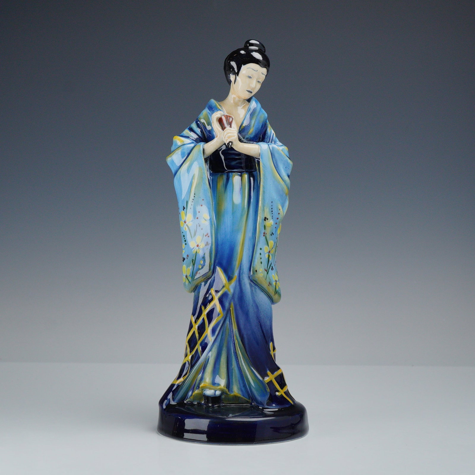 Royal Doulton Porcelain Prototype Figurine with Blue and Yellow Accents, The Geisha: This Royal Doulton porcelain figure is identified as The Geisha. The standing figure is depicted in traditional Japanese attire, featuring a kimono in various shades of blue. The garment is decorated 