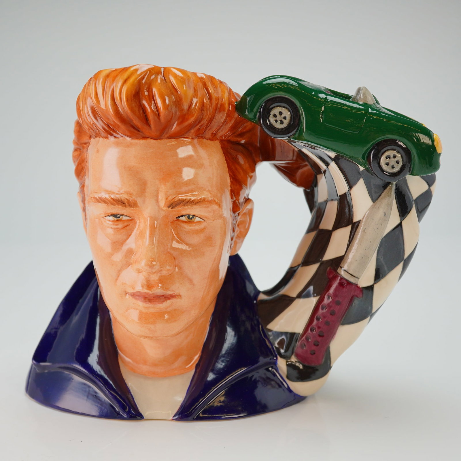 Royal Doulton Prototype Character Jug, James Dean: The piece is themed after the film "Rebel Without a Cause" and features a figure in a dark blue jacket. The handle is composed of three distinct elements: a racing car, a knife, and a checkered flag. 