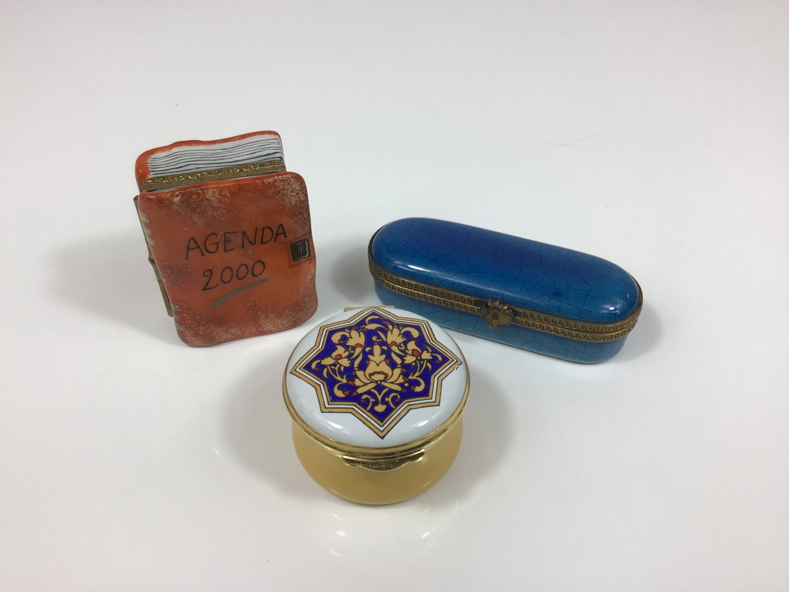Three Limoges Agenda 2000 and Crummles and Co. Trinket Boxes: This lot features three vintage trinket boxes of various origins and materials. The first is a Limoges porcelain box designed in the form of a miniature book. It features a hand-painted orange cover w