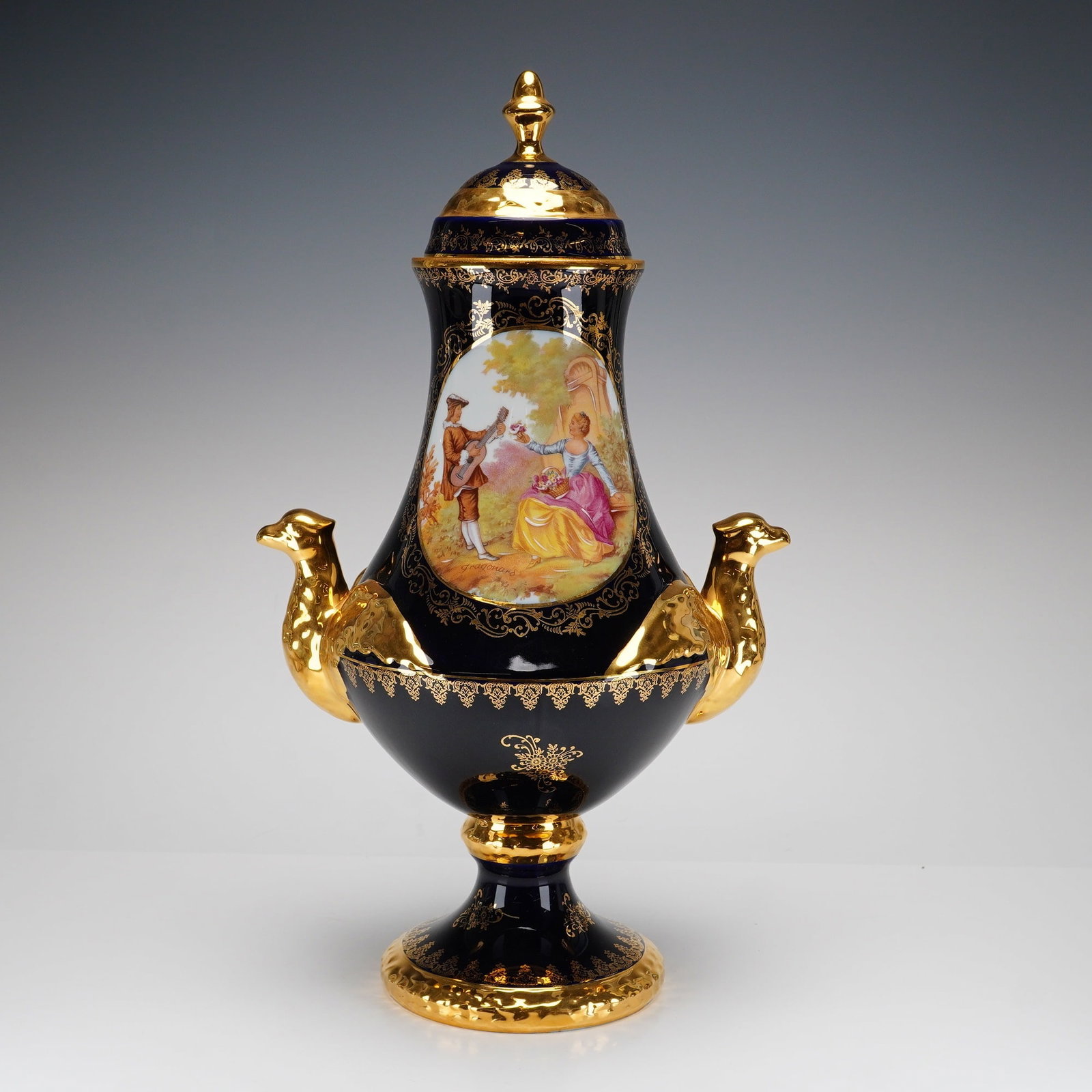 Lemaire Decorateur Limoges Cobalt and Gilt Covered Urn, Courting Couple: This lidded porcelain urn was produced in Limoges, France by Lemaire Decorateur. The piece has a tall, tapering neck, a rounded, bulbous body, and a spreading pedestal foot, all finished in a rich cob