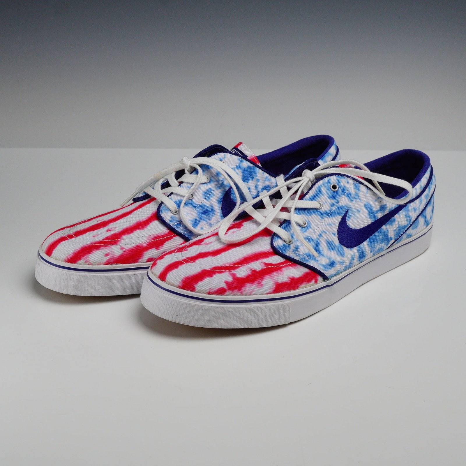 Pair of Nike SB Zoom Stefan Janoski PRM Independence Day Tie-Dye Sneakers, Size 12: A pair of Nike SB Zoom Stefan Janoski Premium low-top skateboarding sneakers featuring an Independence Day tie-dye motif. The white canvas uppers are decorated with red tie-dye stripes across the toe 