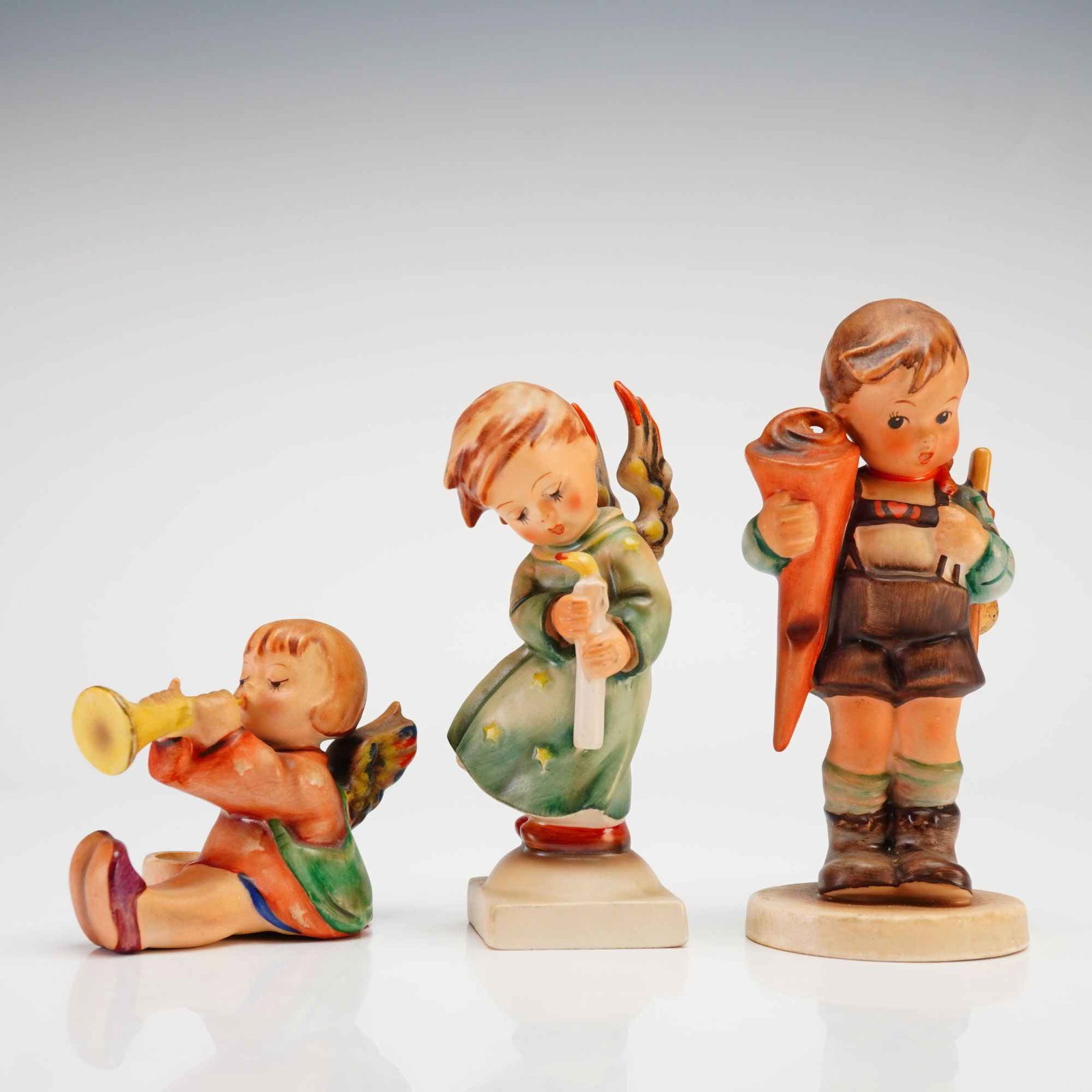 Three Hummel Goebel Figurines Including Joyous News Little Scholar and Heavenly Angel: Three Hummel Goebel hand-painted ceramic figurines comprising the models Joyous News, Little Scholar, and Heavenly Angel. The grouping includes a seated angel playing a trumpet beside an integrated ca