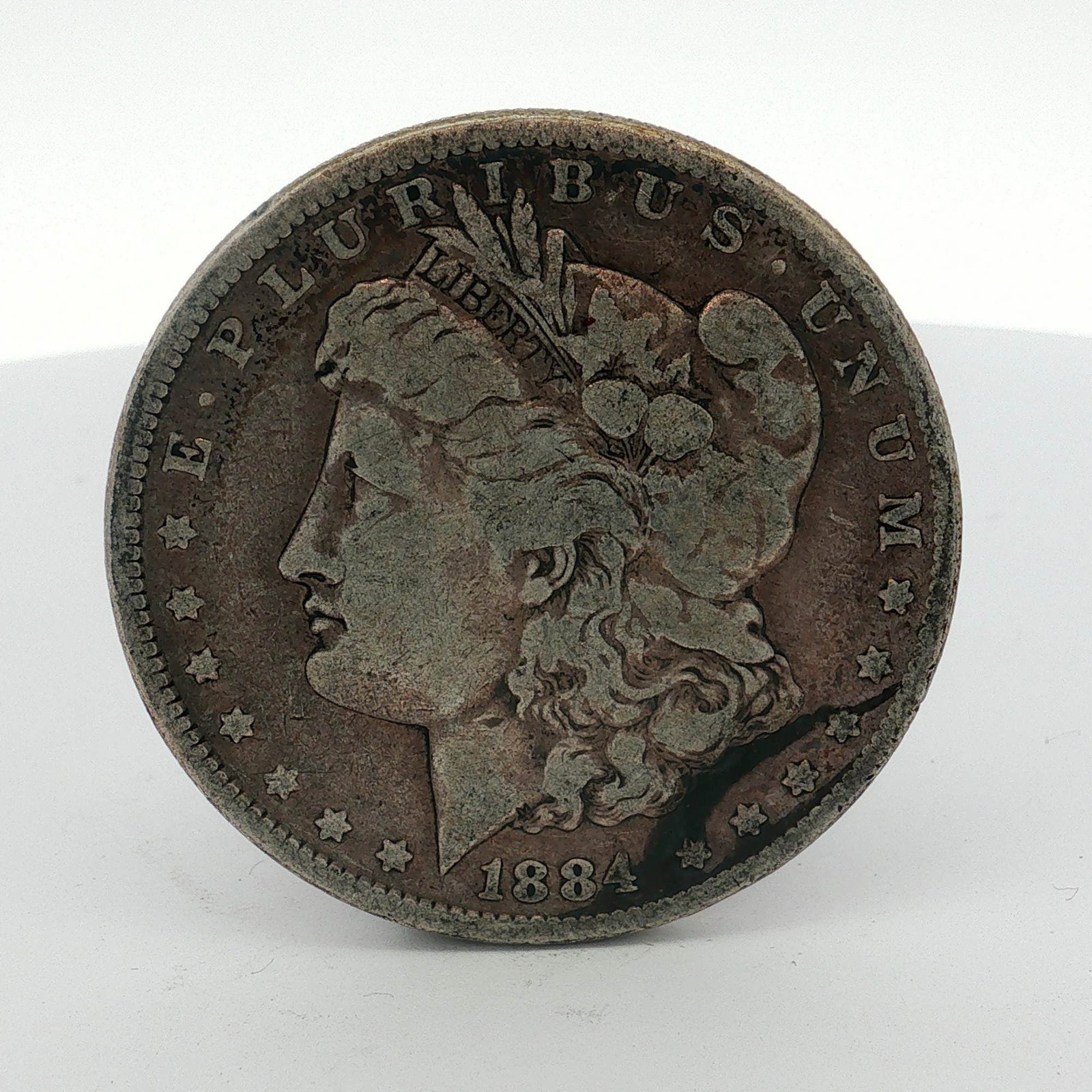 1884 US One Dollar Morgan Silver Dollar, Circulated: This Morgan dollar is a US 1884 silver coin struck at the Philadelphia mint. It is composed of 90% silver and 10% copper with a fineness of .900 silver. The obverse features the Liberty Head design ty