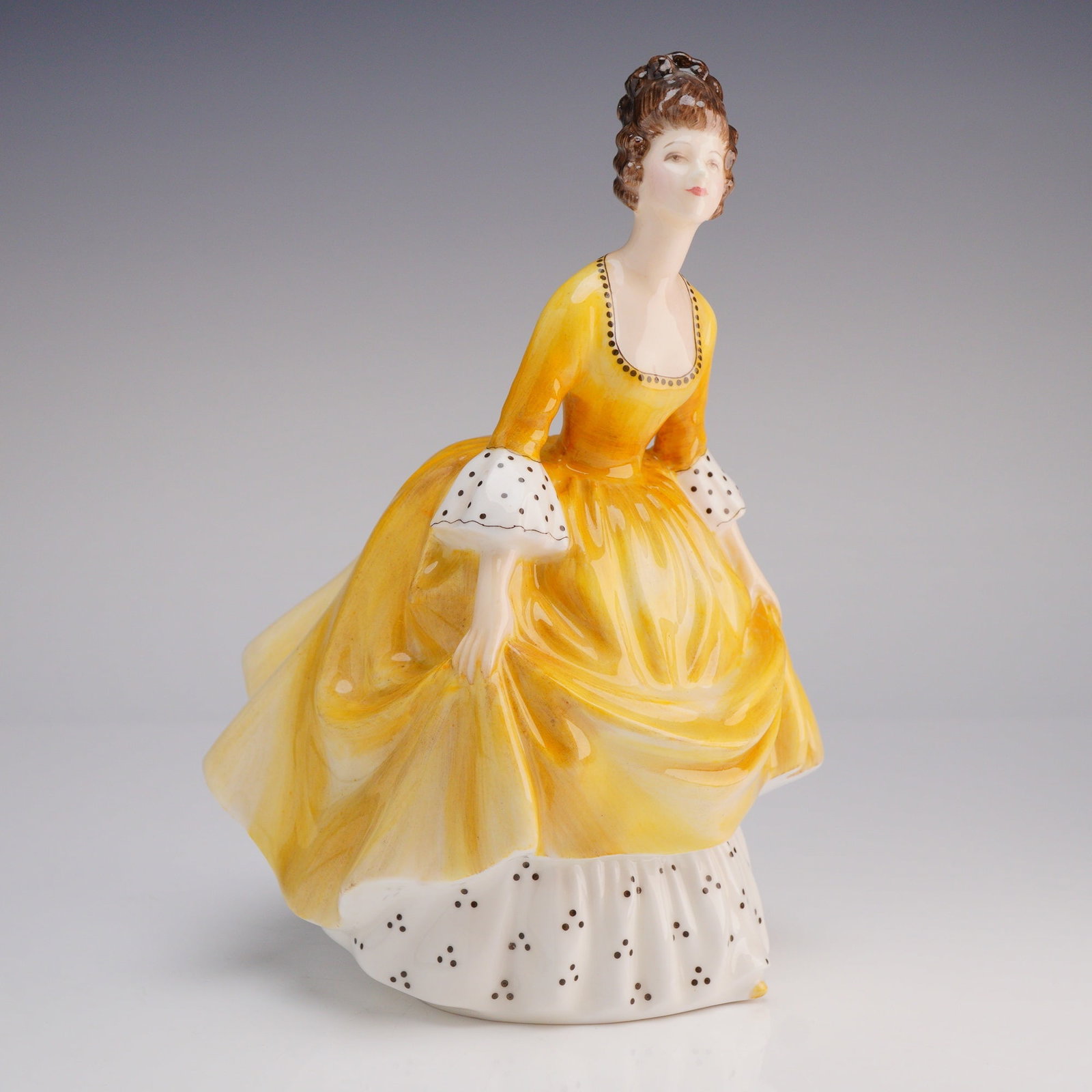 Royal Doulton Coralie Figurine HN2307: The figure depicts a standing woman in a flowing, high-gloss yellow gown. The design is accented with white and black polka dot detailing visible on the wide underskirt and the puffed sleeve cuffs. Th