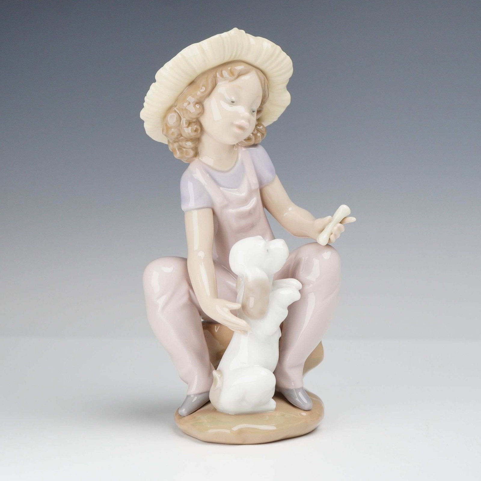 Lladro Porcelain Figurine Friends Forever 01006680: The piece depicts a young girl with curly hair wearing a wide-brimmed straw hat, a lavender short-sleeved shirt, and pink overalls. She is seated on a rock and looks down at a small white dog jumping 