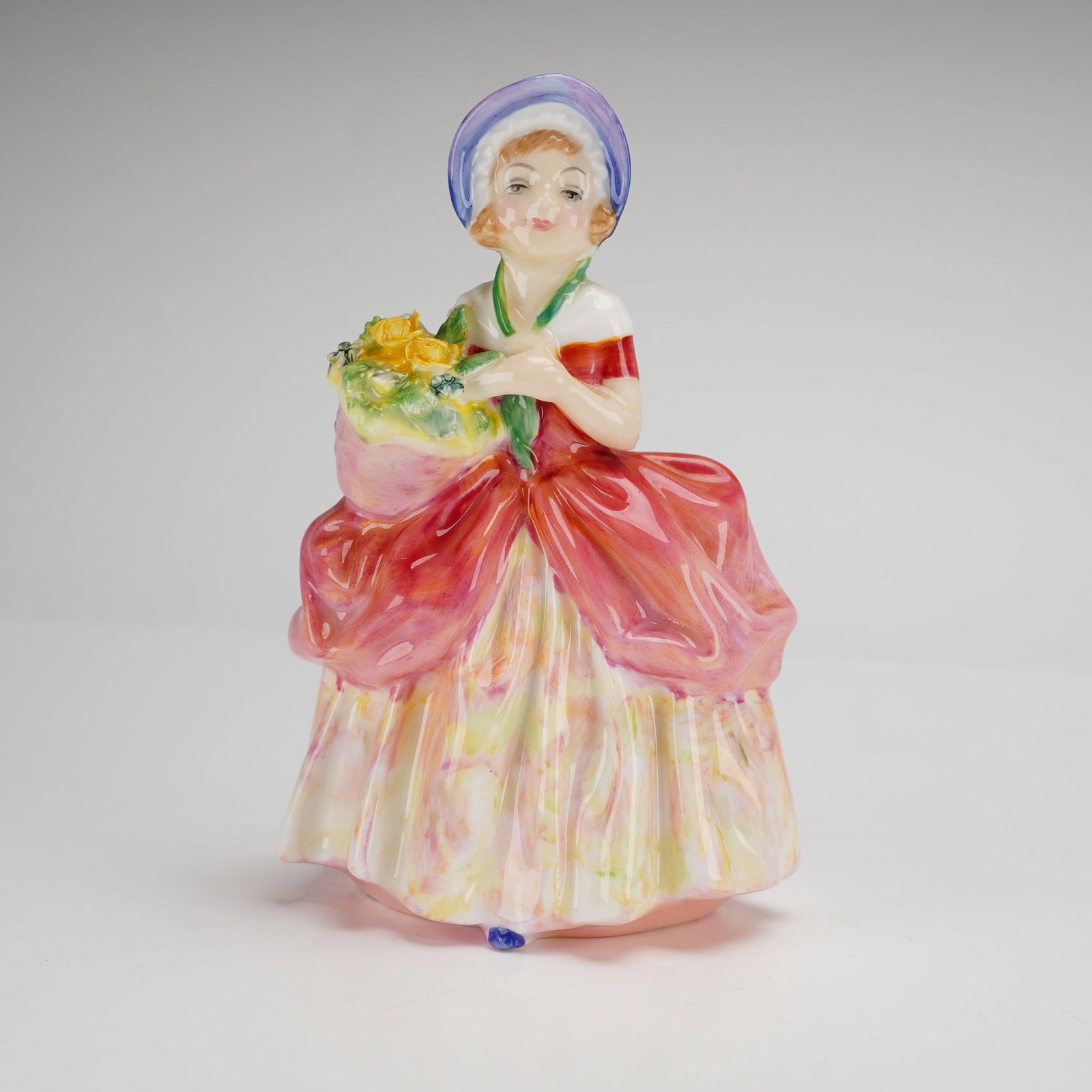 Royal Doulton Bone China Figurine Cissie HN1809: This Royal Doulton bone china figurine, titled Cissie (HN1809), was designed by Leslie Harradine. The figure depicts a young woman wearing a blue and purple bonnet and a dress with a voluminous red ov