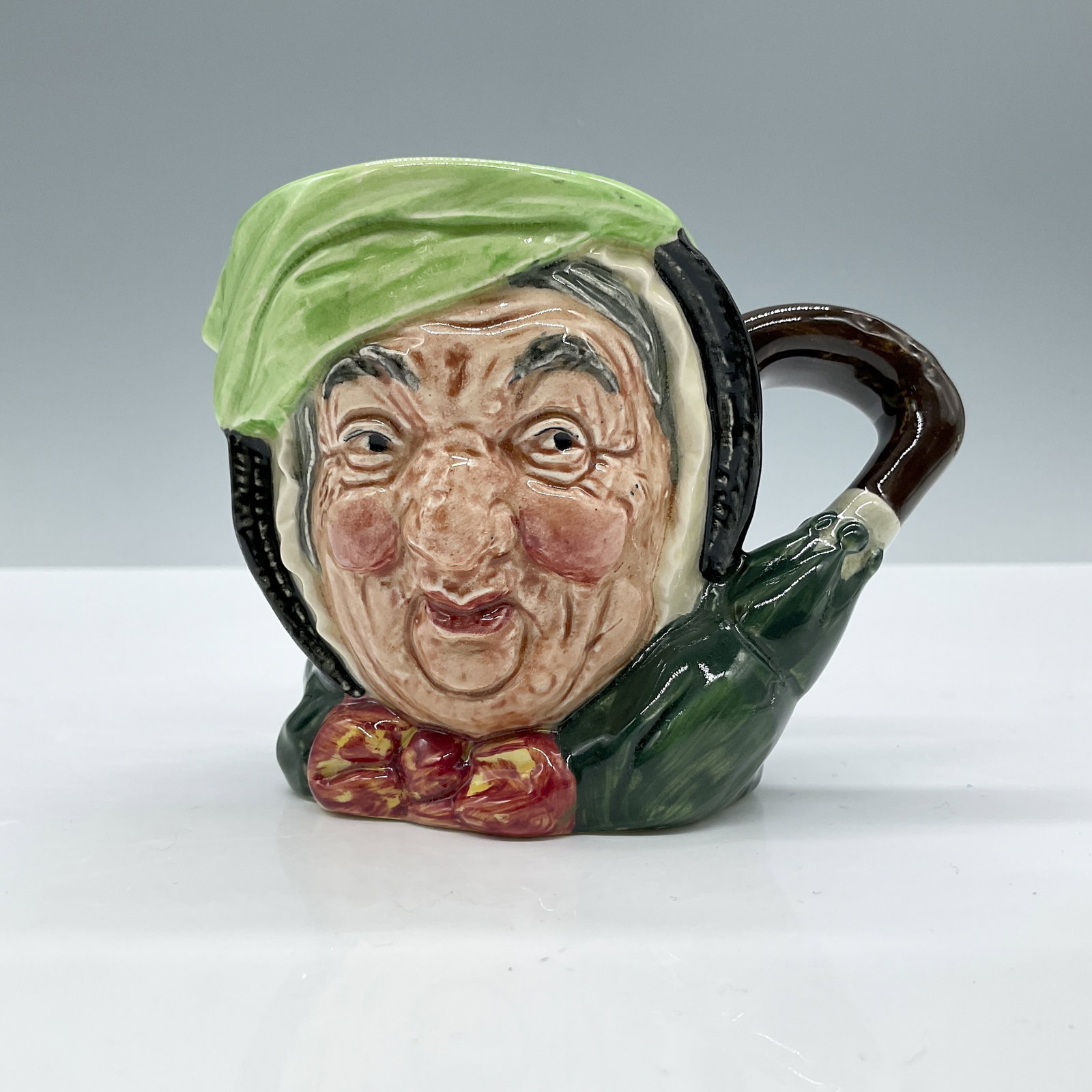 Sairey Gamp D5528 - Small - Royal Doulton Character Jug: Black hair; light green band; dark green coat; yellow and burgundy bow; green umbrella with brown handle. Royal Doulton backstamp. Artist: Leslie Harradine and Harry Fenton Dimensions: 3.25"H Date or 