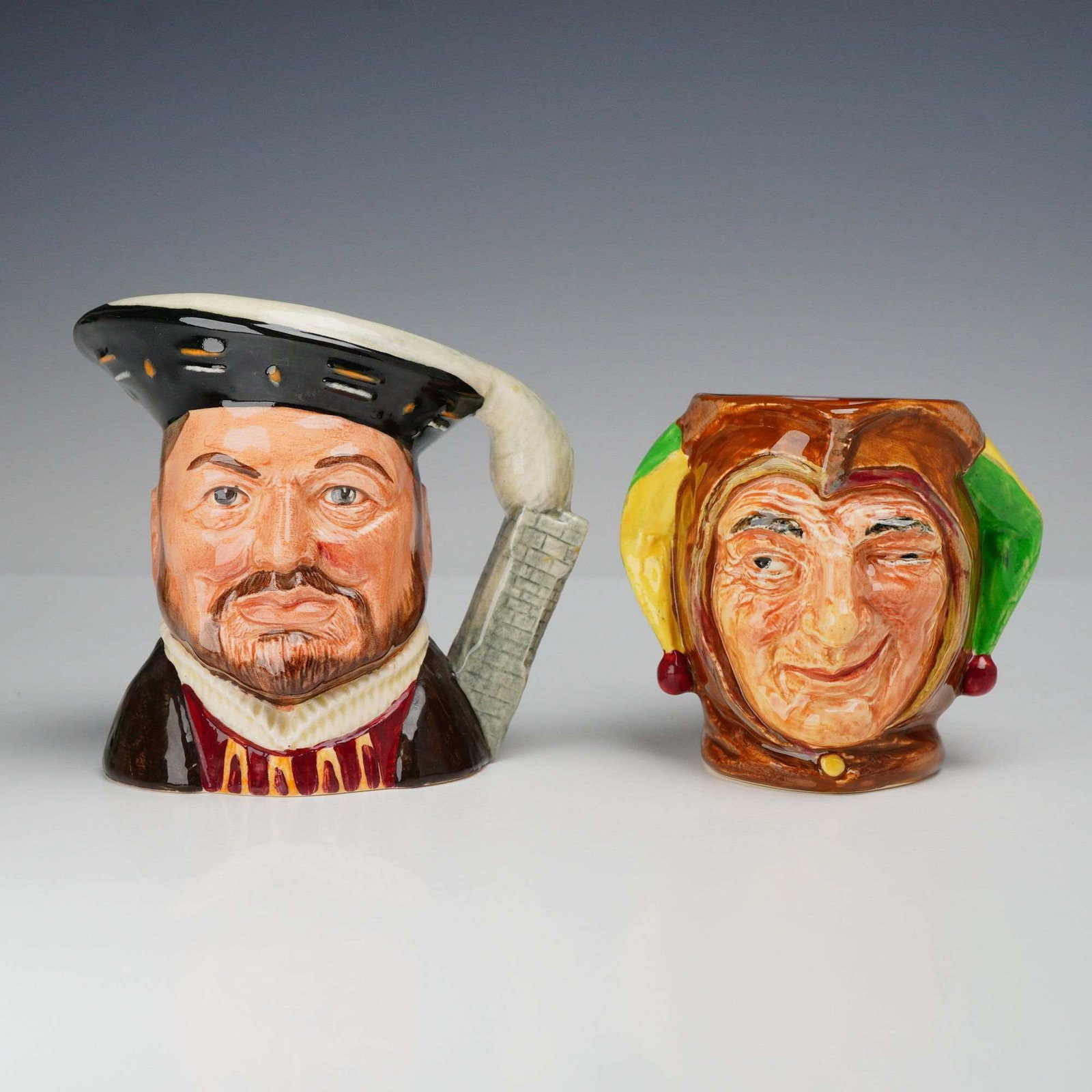 2pc Royal Doulton Character Jugs, Henry VIII D6647 and Jester D5556: This lot consists of two Royal Doulton glazed earthenware character jugs. The first jug depicts King Henry VIII, model number D 6647. The figure is rendered with a brown beard and mustache, wearing a 