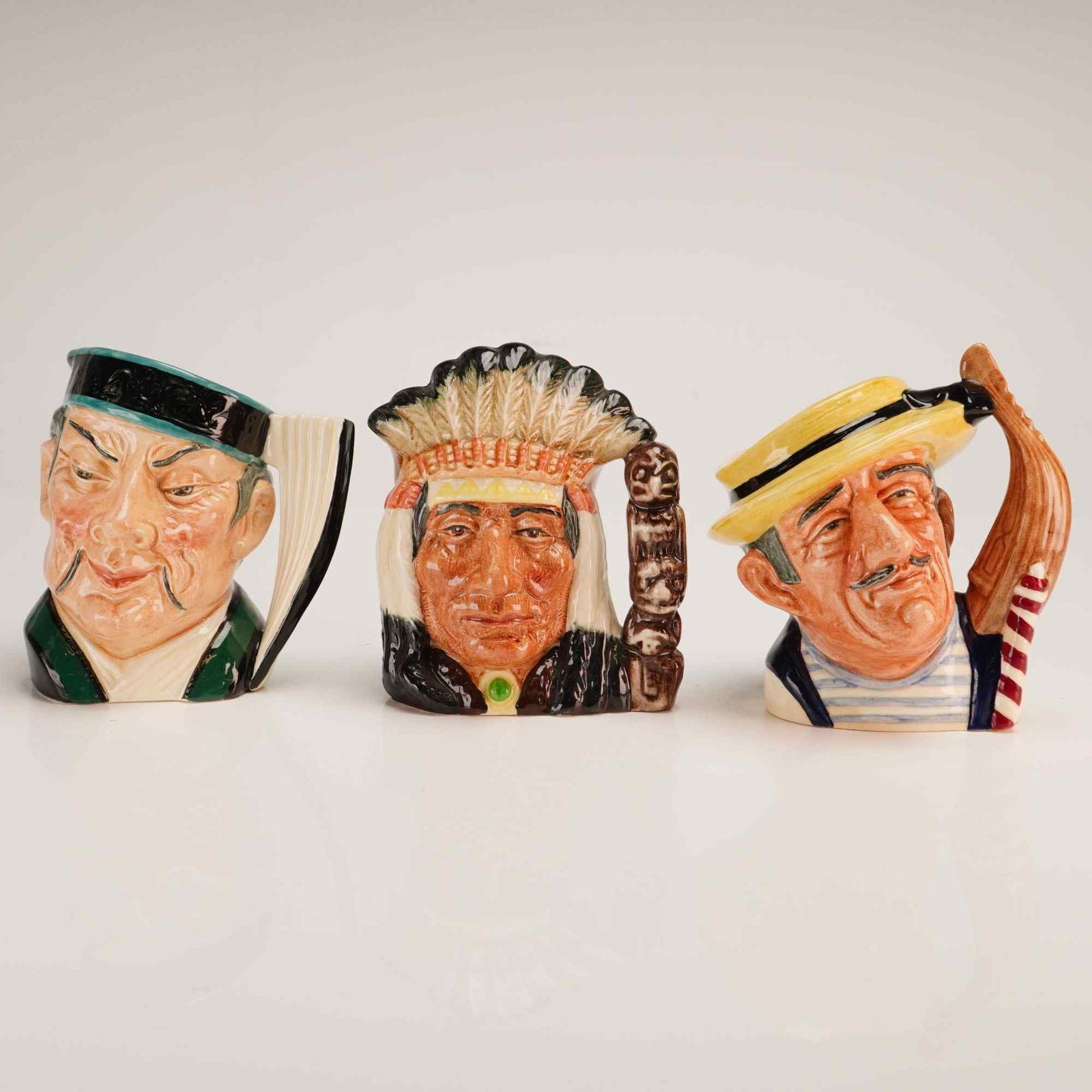 Three Royal Doulton Small Character Jugs- The Mikado D6502, North American Indian 6614, & Gondolier: This collection consists of three small-sized glazed earthenware character jugs manufactured by Royal Doulton in England. Each jug features a distinctive molded form with hand-painted polychrome decor