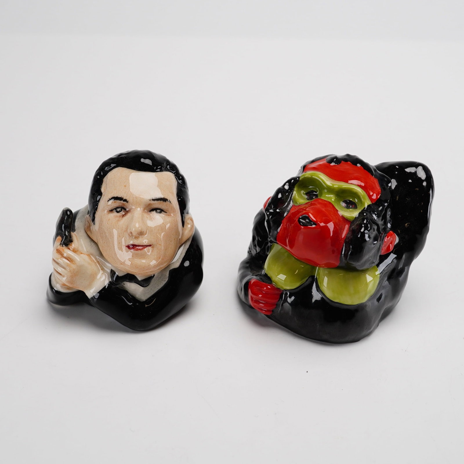 Lot of 2 Kevin Francis Porcelain Face Pots - James Dink and Clyde the Chimpanzee: The set includes two specific character studies: James Dink and Clyde the chimpanzee. The James Dink container depicts a man in a black tuxedo and bow tie holding a martini glass. The interior of the 