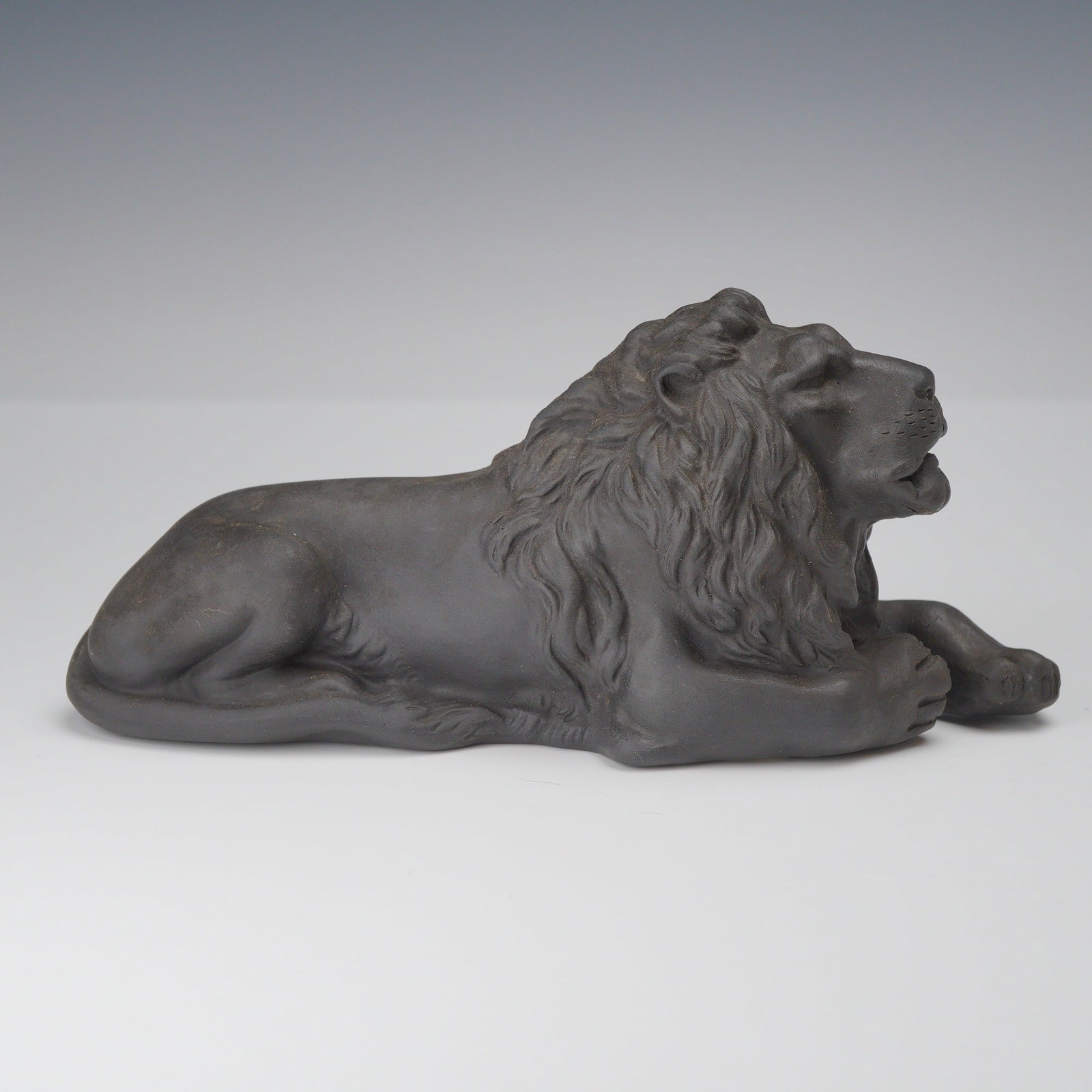 Wedgwood Black Basalt Reclining Lion Figurine: This is a Wedgwood black basalt figurine depicting a lion in a reclining or recumbent pose. The piece is crafted from fine-grained, unglazed black stoneware with a matte finish. The lion is stylized w