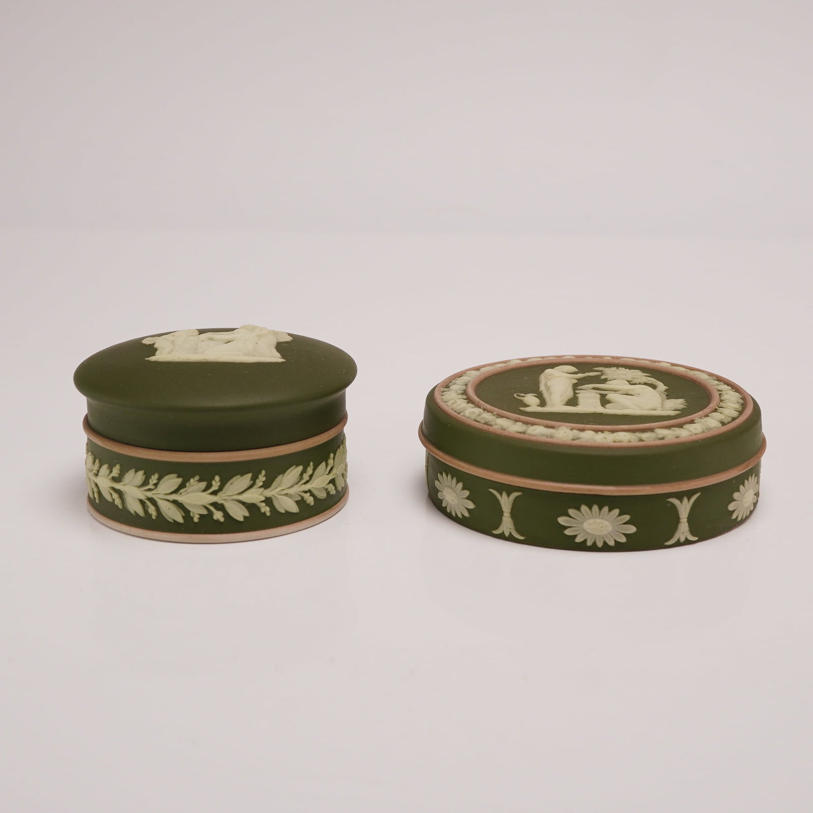Two Wedgwood Olive Green Jasperware Lidded Boxes from the Rubin Collection: This lot consists of two circular lidded boxes produced in Wedgwood jasperware. These matte-finish stoneware items feature an olive green ground decorated with white sprigged neoclassical relief and t