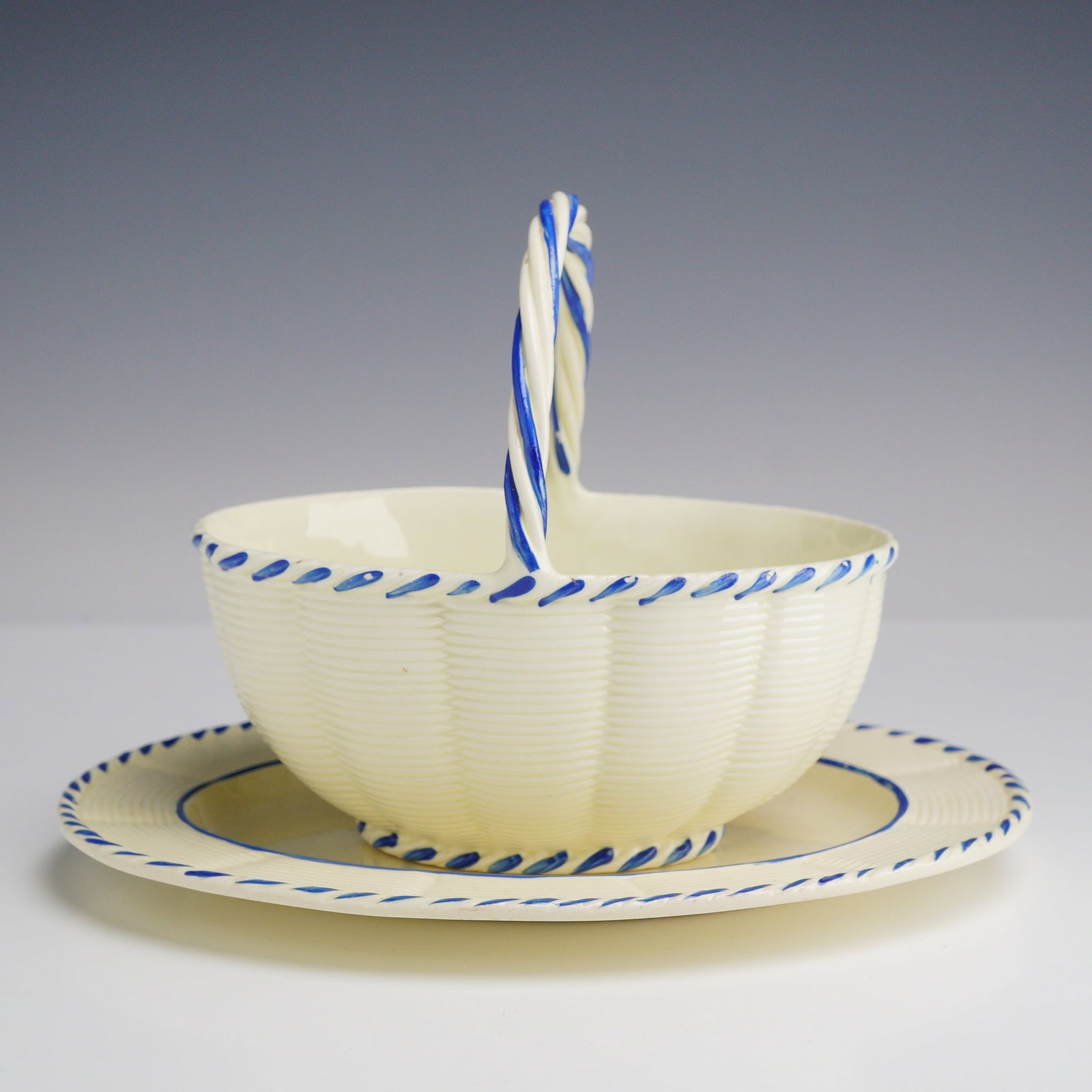 Wedgwood Creamware Basket and Plate: This set consists of an oval creamware basket with a twisted overhead handle and a matching oval plate by Wedgwood. Both pieces are made from cream-colored earthenware. The basket is molded with a bas