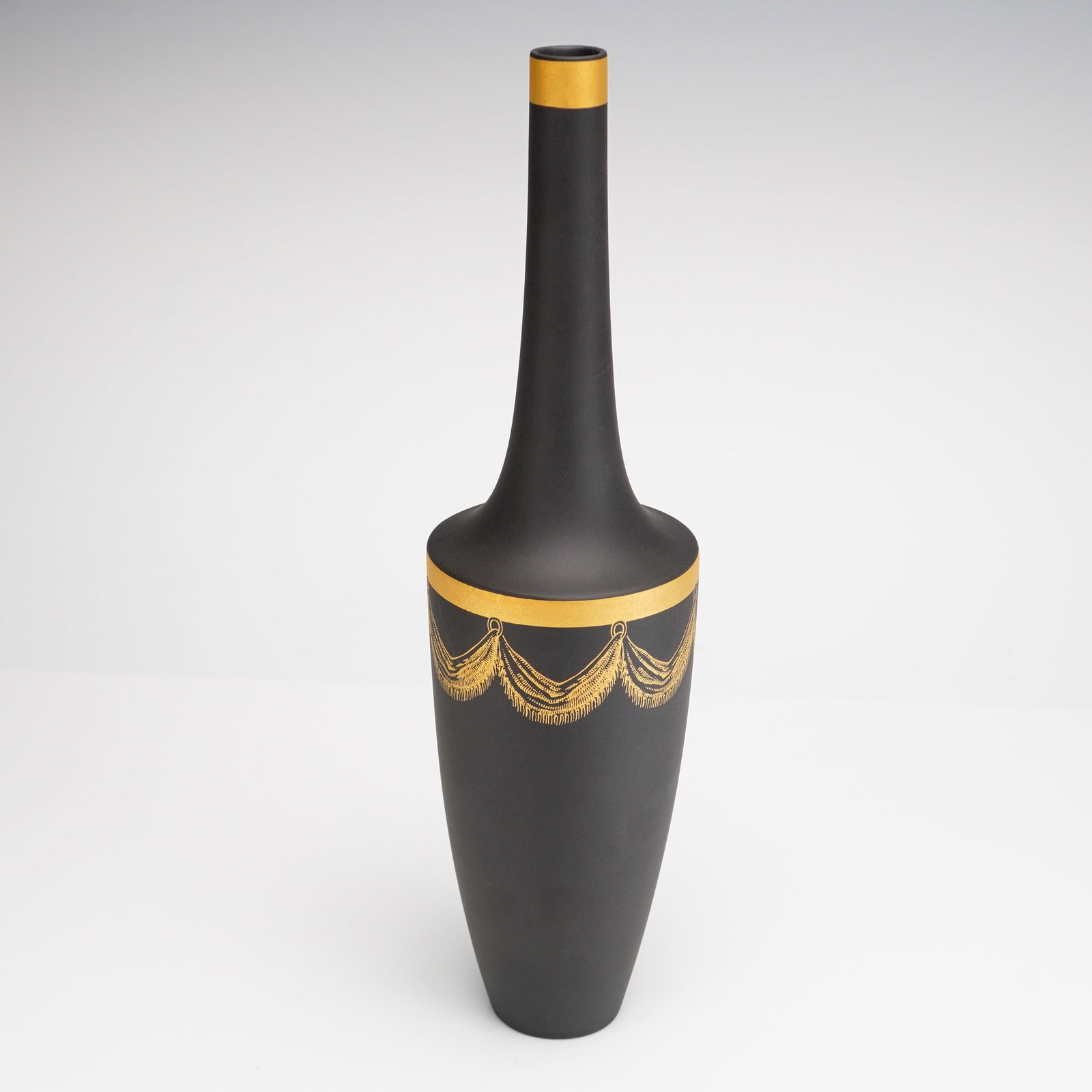 Wedgwood Black Jasperware Gilded Vase: This slender Wedgwood black jasperware vase features a matte-finished stoneware body. The object is designed in a tall bottle form characterized by a narrow straight neck, a high flared shoulder, and 