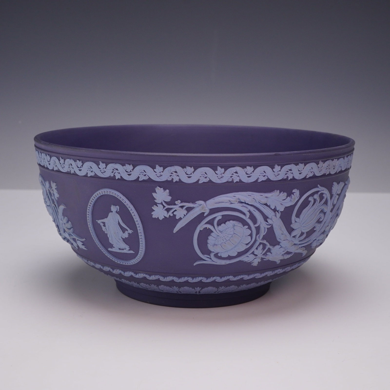 Wedgwood Lilac Jasperware Arabesque Bowl: A Wedgwood lilac jasperware footed bowl in the Arabesque pattern featuring classicizing sprigged relief decoration in white. The exterior body is ornamented with two oval medallions containing classic