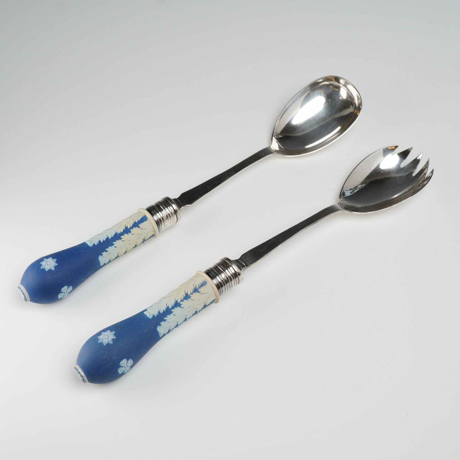 A Pair of Wedgwood Blue Jasperware and Silver-Plated Salad Servers: A pair of salad serving utensils comprising a large spoon and a three-tined fork. These pieces feature cobalt blue jasperware ceramic handles decorated with white bas-relief sprigging in acanthus leaf