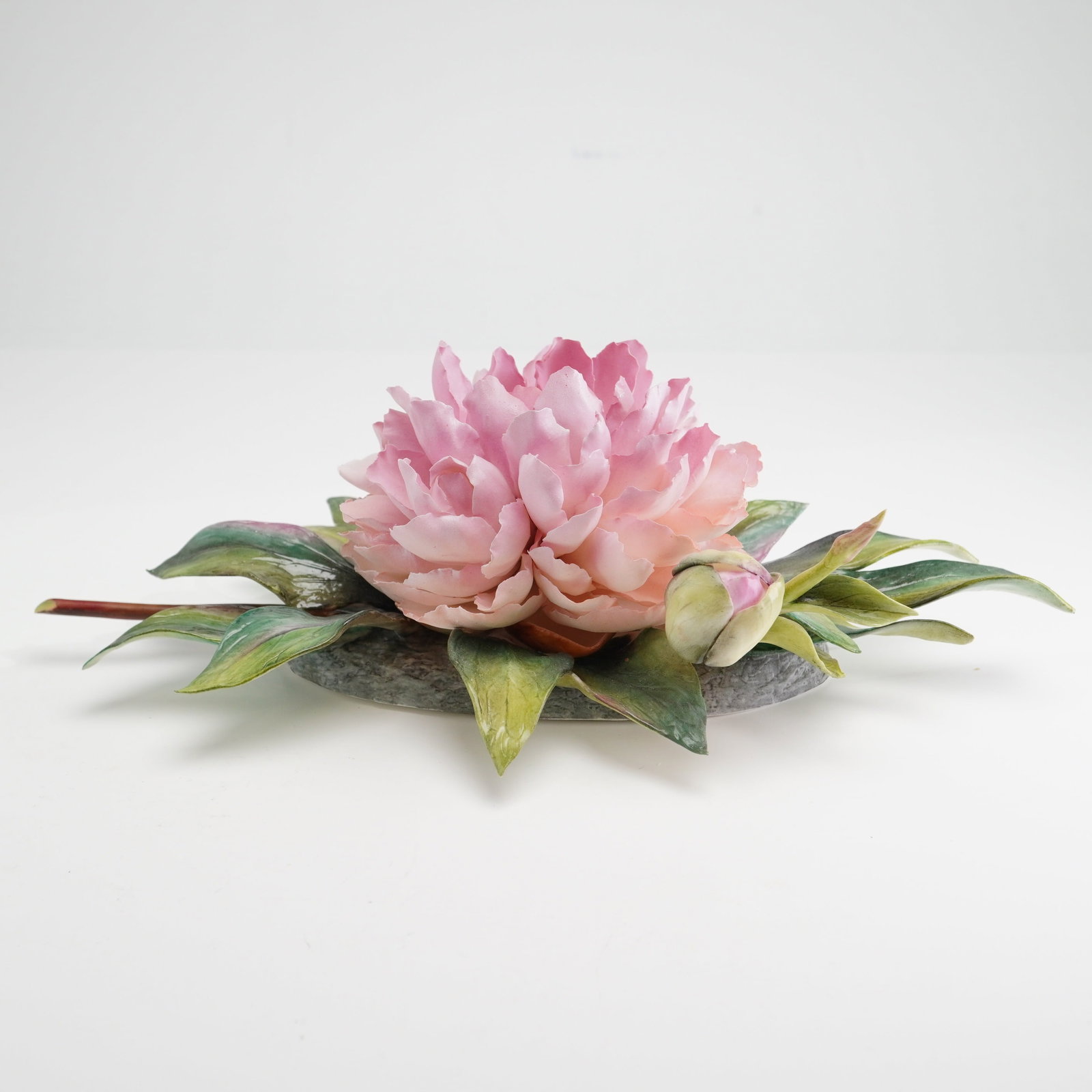 Boehm Bone Porcelain Sculpture, Pink Peony: A bone porcelain sculpture modeled as a fully bloomed pink peony and bud on stem with detailed painted leaves. The underside of the base features the Boehm backstamp. Dimensions: 11"L x 7"W x 3.75"H M