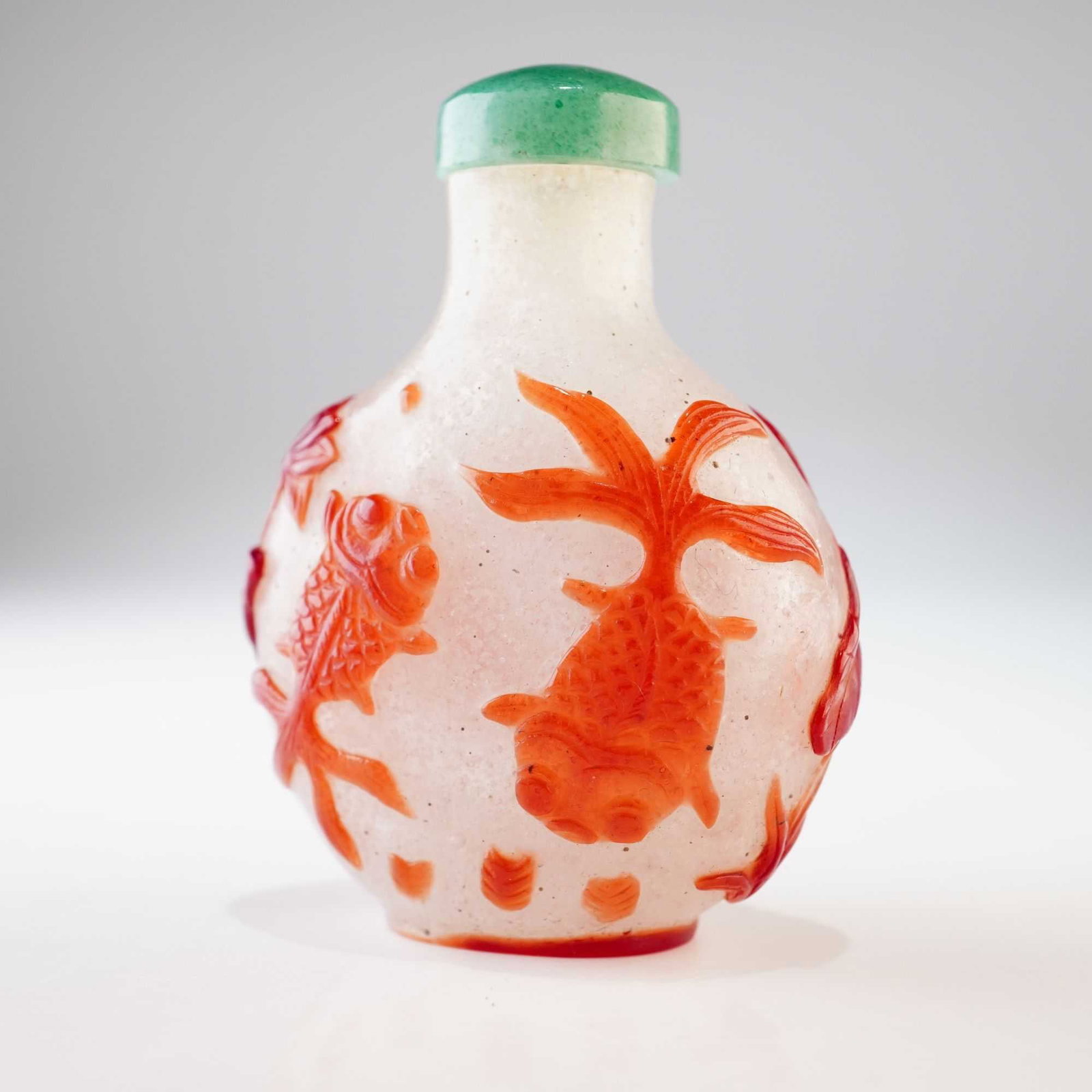 Peking Glass Snuff Bottle, Brown Overlay Goldfish and Lotus: A clear round snowflake Peking glass snuff bottle decorated with brown relief overlay showcasing goldfish, lotus flowers and leaves. It is topped with a green stopper. Dimensions: 1.25"L x 1"W x 2.25"