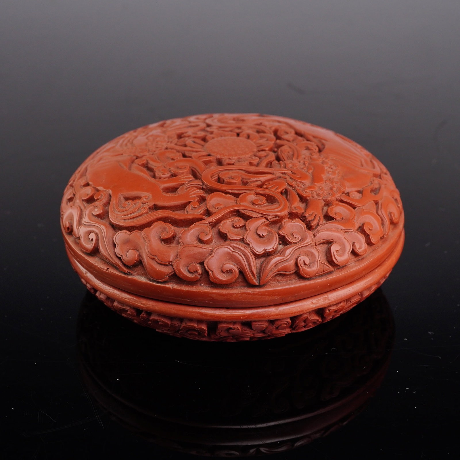 Chinese Carved Cinnabar Lacquer Foo Dog Jewelry Box: A circular enameled box depicting two foo dogs on the lid. The piece is constructed of cinnabar lacquer over a composite core, featuring deep relief carvings. The central scene on the lid illustrates 