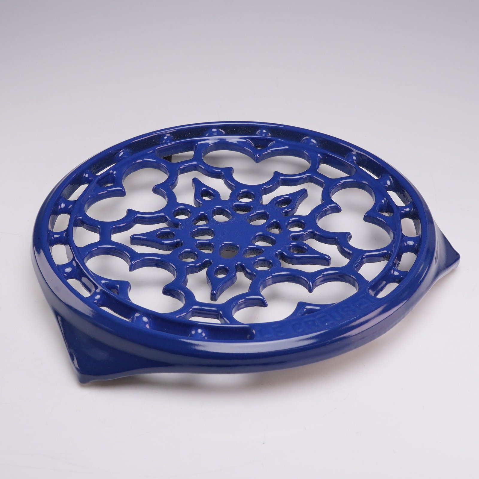 Le Creuset Cobalt Blue Enameled Cast Iron Deluxe Round Trivet: A Le Creuset deluxe round trivet constructed from enameled cast iron in a vibrant cobalt blue finish. The circular piece features an intricate openwork filigree design consisting of symmetrical geomet