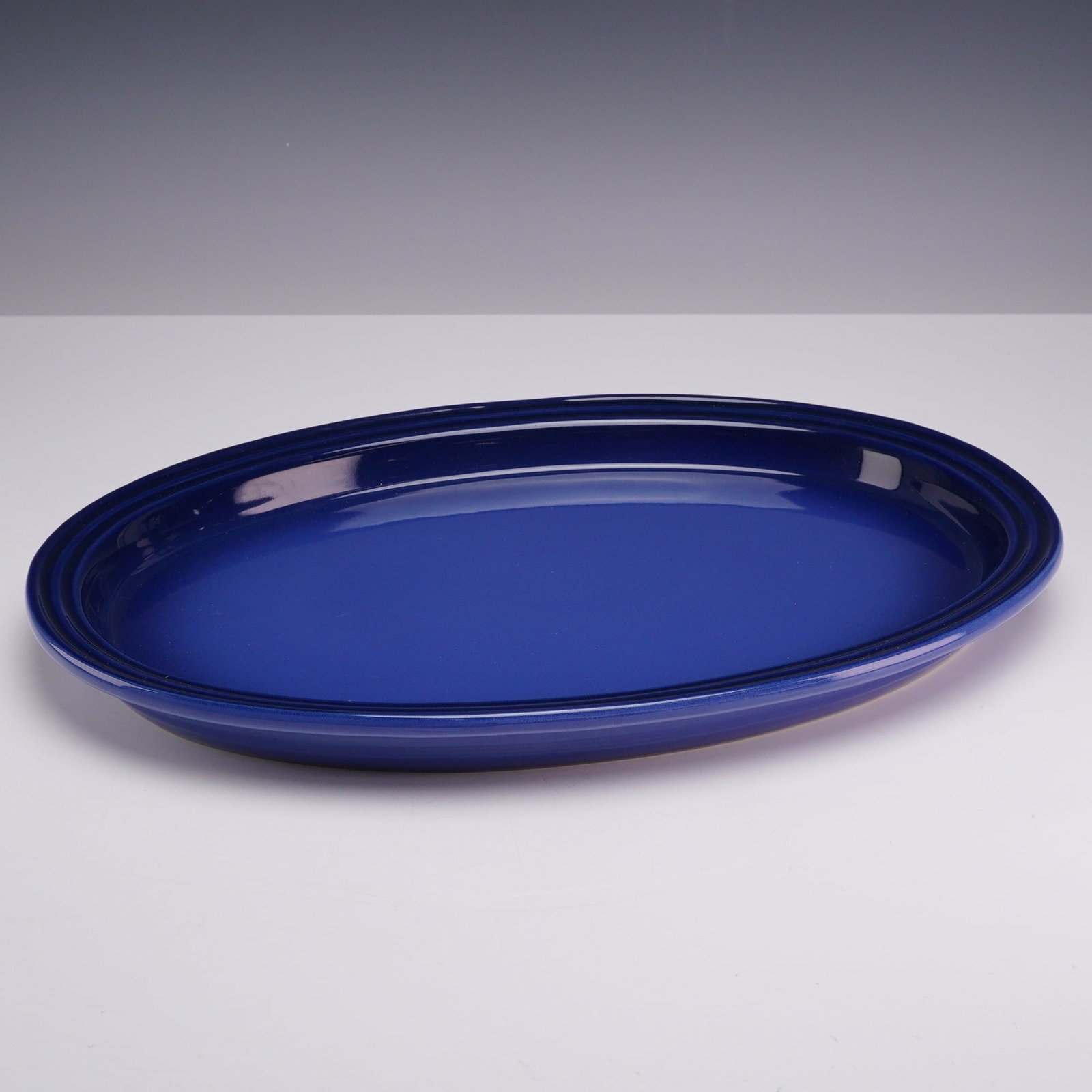Le Creuset Cobalt Blue Enameled Cast Iron Oval Platter: A Le Creuset cobalt blue enameled cast iron oval serving platter, featuring a deep blue high-gloss finish. The design includes a wide rim with the maker's signature concentric triple-ring motif. The i