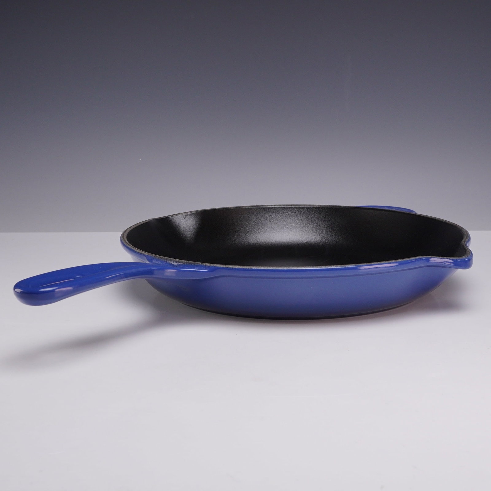 Le Creuset Cobalt Blue Enameled Cast Iron Skillet: A Le Creuset enameled cast iron skillet featuring a vibrant cobalt blue exterior finish and a black satin interior enamel. The vessel is designed with a traditional long handle and an integrated helpe