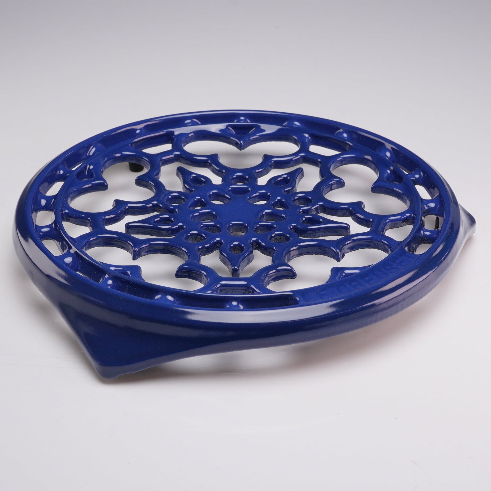 Le Creuset Enameled Cast Iron 9-Inch Deluxe Round Trivet in Cobalt Blue: A circular footed trivet manufactured by Le Creuset, crafted from cast iron and finished with a cobalt blue vitreous enamel. The design features an intricate openwork filigree pattern with a central s