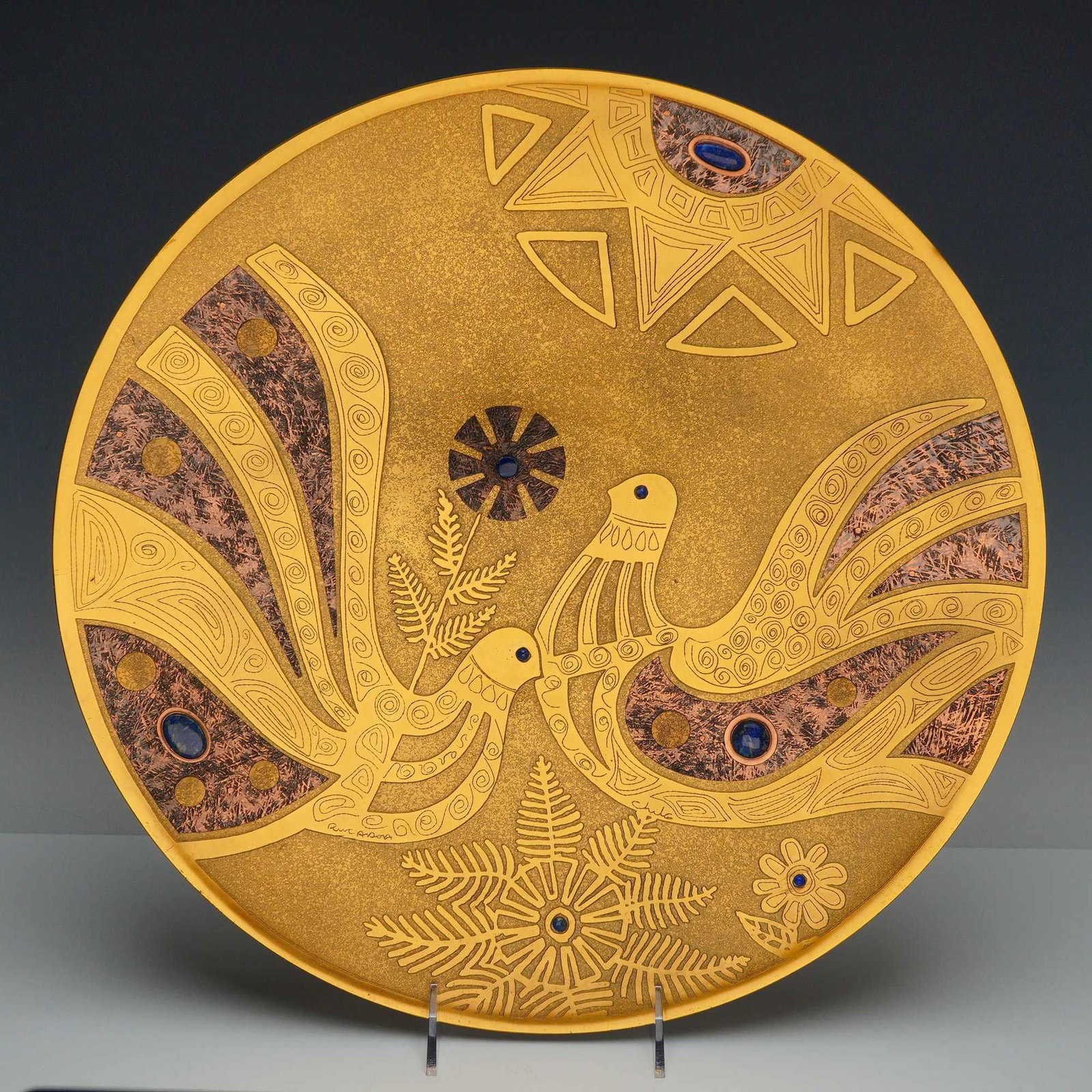 Ruandora Chilean Mixed-Metal Brass and Copper Wall Plate with Lapis Lazuli: This decorative Chilean mixed-metal charger is a work by the maker Ruandora, featuring a modernist composition of two birds amidst stylized floral and fern-like foliage. The circular plate is construc
