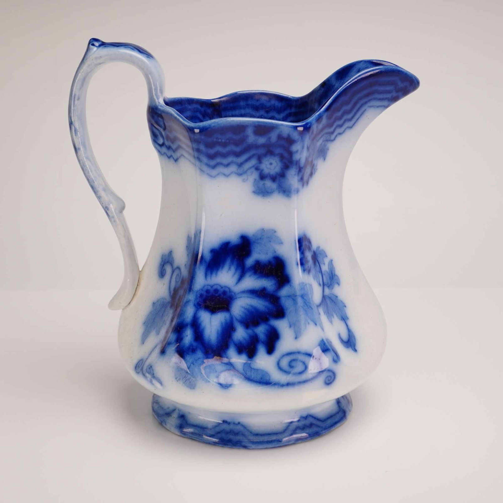 Antique Flow Blue Transferware Ironstone Water Pitcher, Late 19th Century: This flow blue transferware pitcher is composed of white ironstone or earthenware. The vessel features a bulbous body that tapers toward a flaring, scalloped rim with a pinched spout. It is equipped