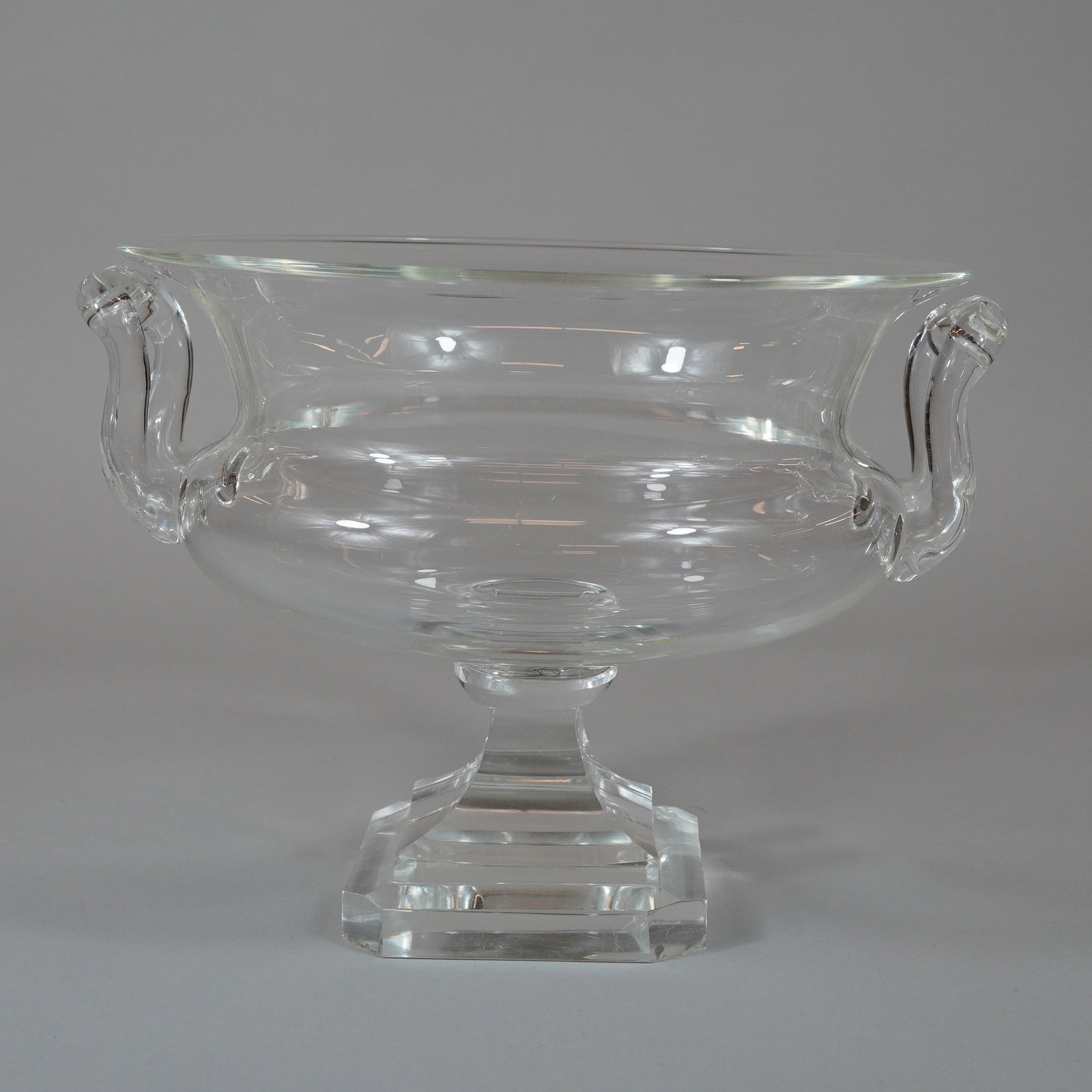 Steuben Crystal Two-Handle Pedestal Centerpiece Bowl: A large colorless crystal centerpiece bowl of urn form produced by Steuben. The piece features a wide flared rim above a bulbous body, flanked by two applied scrolled handles. The bowl is supported by