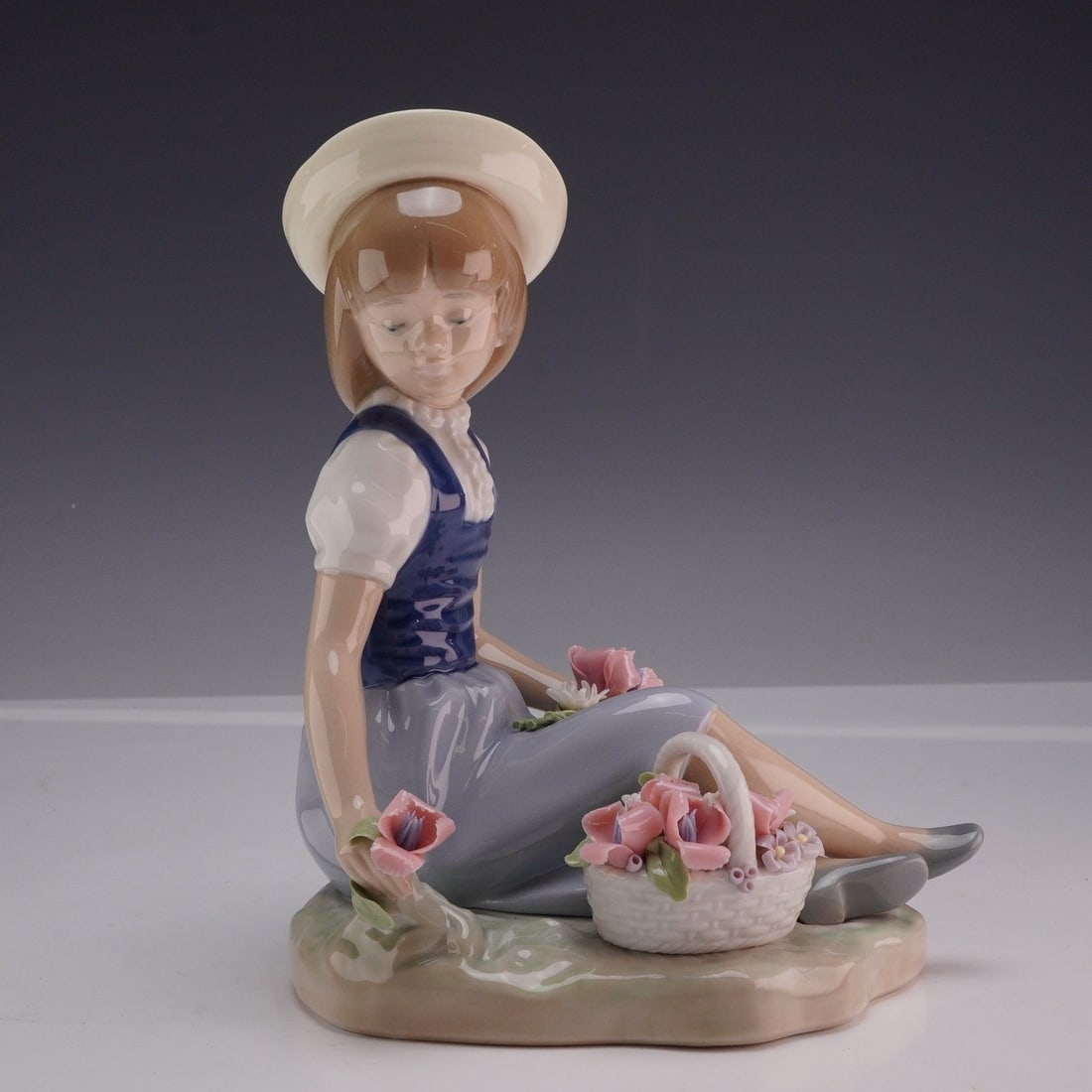 Lladro Porcelain Figurine Picking Flowers 01001287 with Original Box: This piece was designed by sculptor Juan Huerta and depicts a young girl in a seated, pensive pose on a grassy, earth-toned base. The figure is dressed in a wide-brimmed cream hat, a white short-sleev