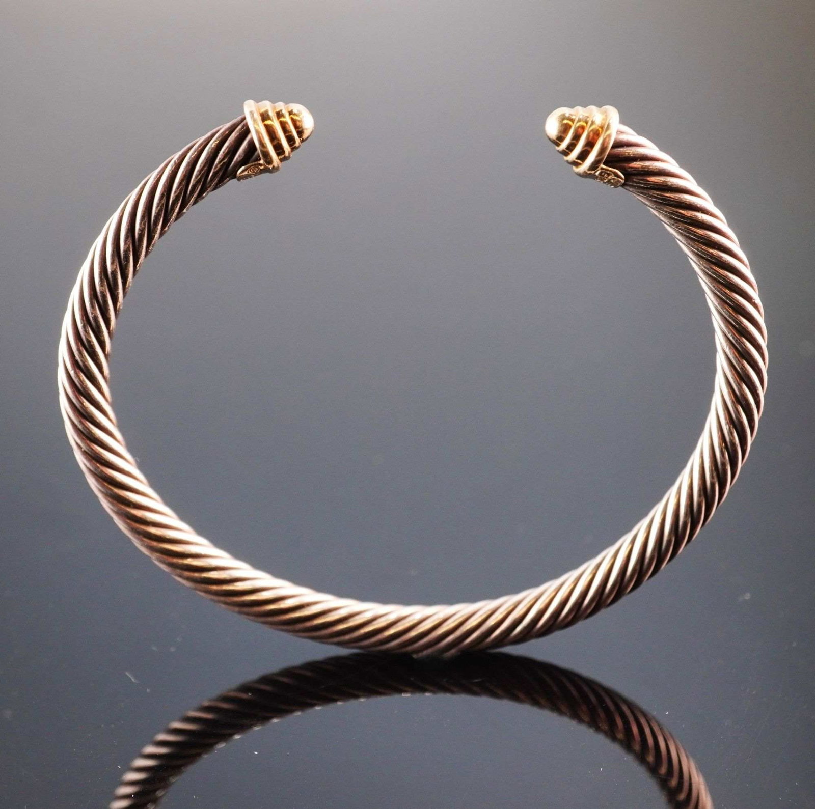 David Yurman Sterling Silver and 14K Yellow Gold 5mm Cable Cuff Bracelet: A designer cable cuff bracelet by David Yurman featuring the signature 5mm twisted cable band. The bracelet is constructed of sterling silver with two ribbed conical finials crafted in 14 karat yellow