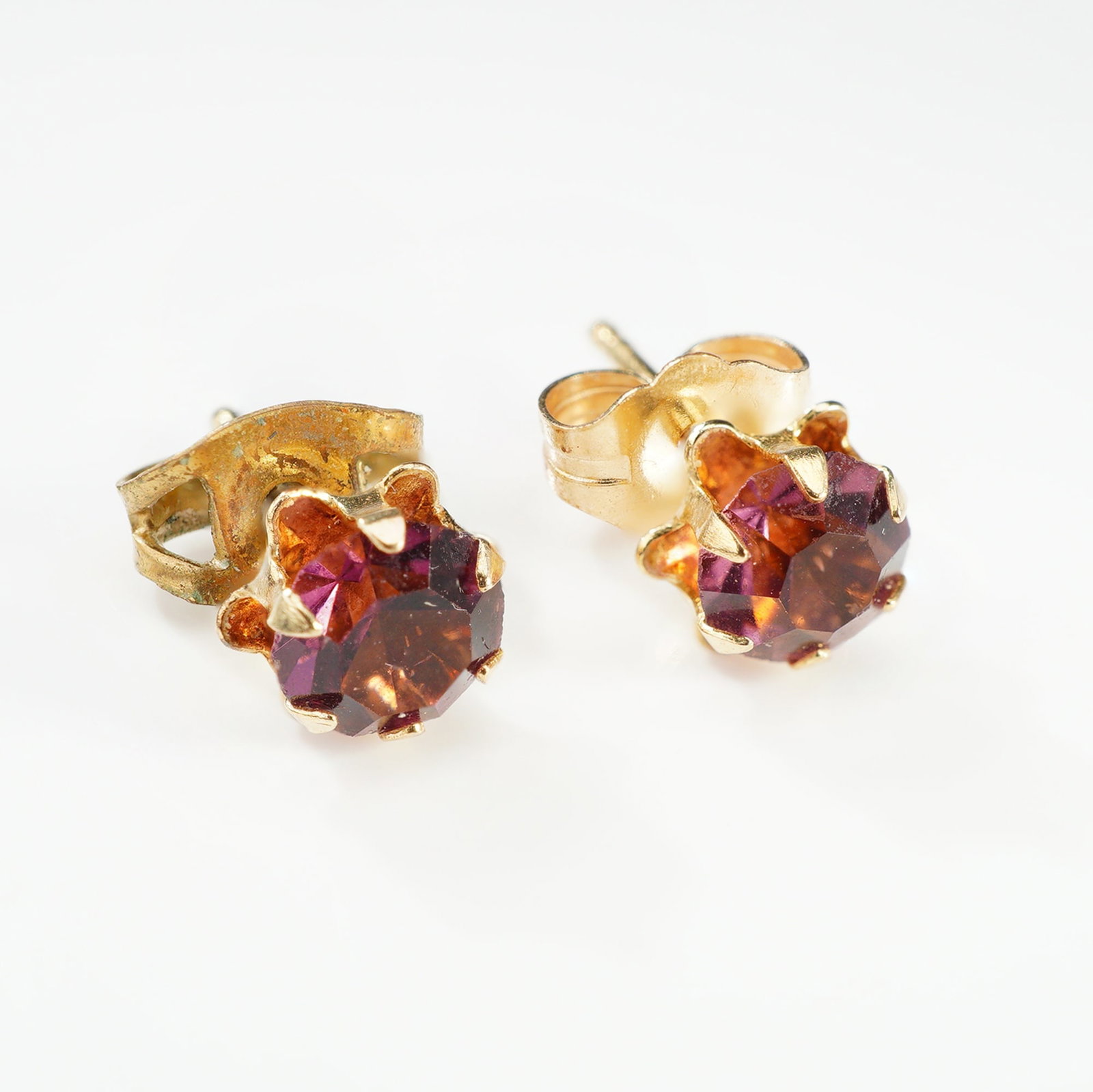 Pair of 14KT Yellow Gold and Amethyst Stud Earrings: This is a matched pair of vintage stud earrings featuring round-cut, faceted gemstones of a deep reddish-purple hue. The stones are held in ornate six-prong crown-style basket settings. The earrings a