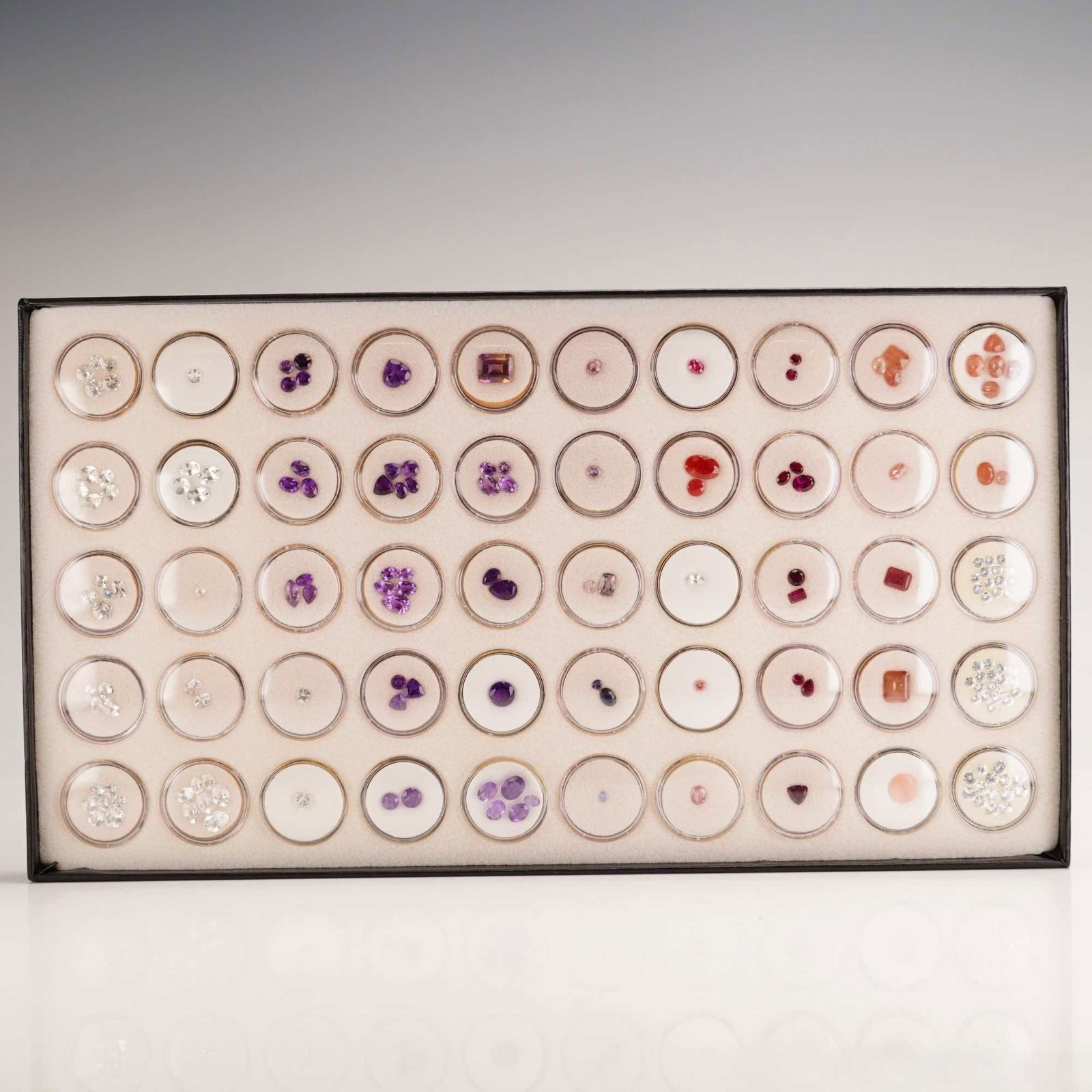 Assortment of Mixed Gemstones, Collectors Group: This assortment of mixed gemstones is presented as a collectors group, housed in a black rectangular display case containing multiple circular clear-lidded gem jars with white foam inserts. The collec
