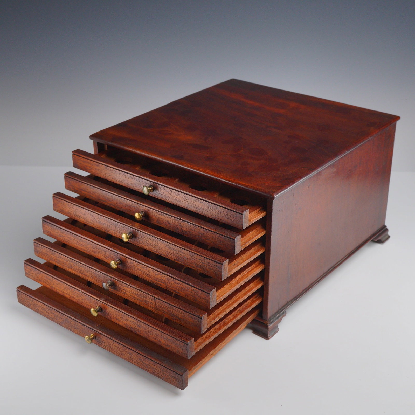 Mahogany Seven-Drawer Coin Cabinet: This mahogany coin cabinet features a rectangular case housing seven shallow drawers. Each drawer front is fitted with a small brass pull knob. The interior of every drawer contains precision-cut circ